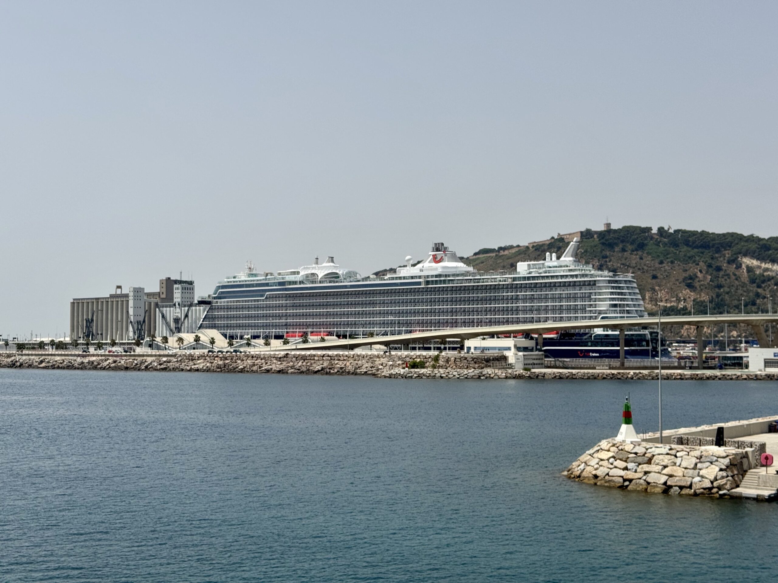Large white cruise ship moored at a harbor with a rocky breakwater and hillside in the background.
