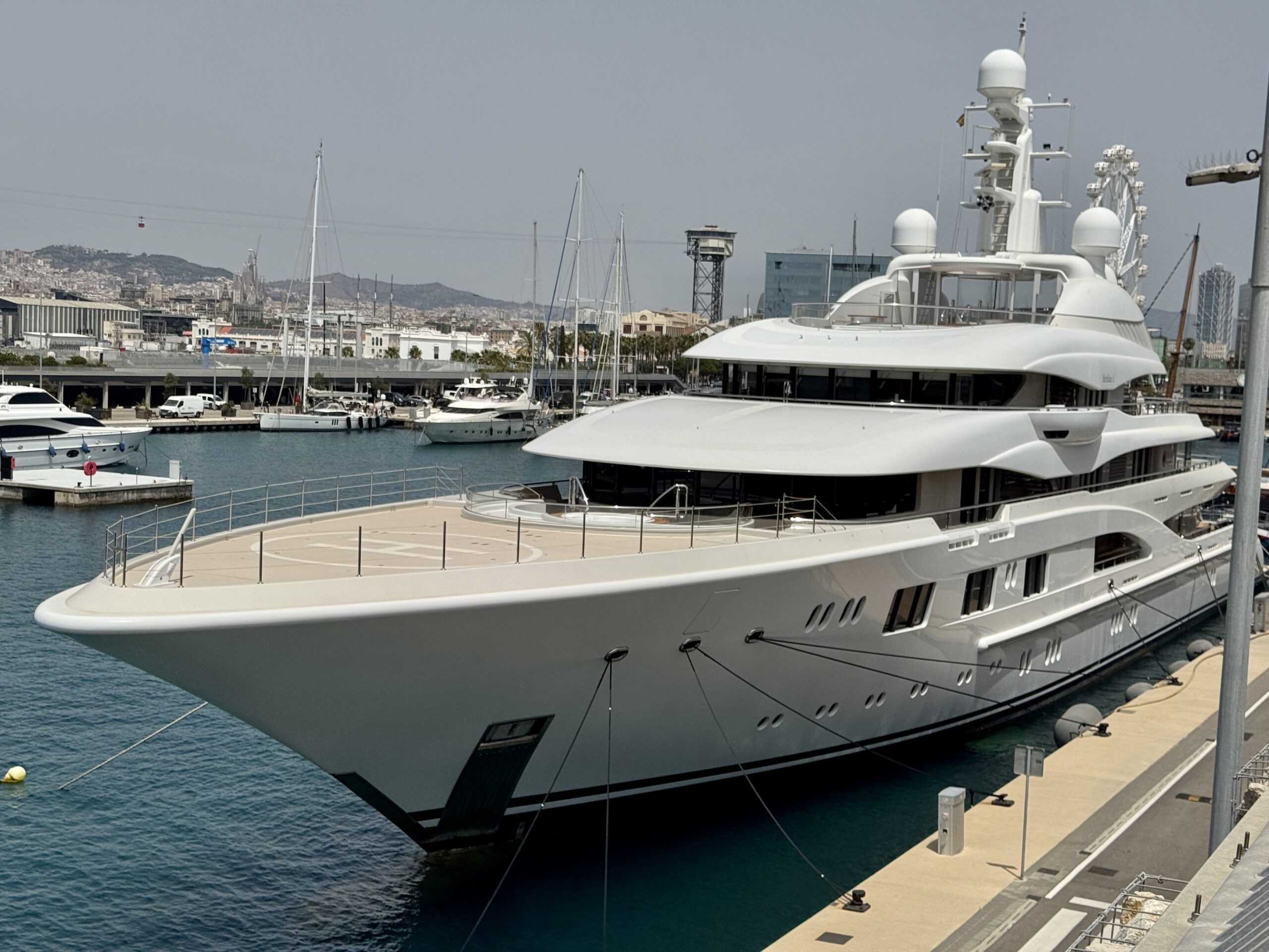 Large white luxury yacht docked at a marina with calm blue water and city skyline in the background.