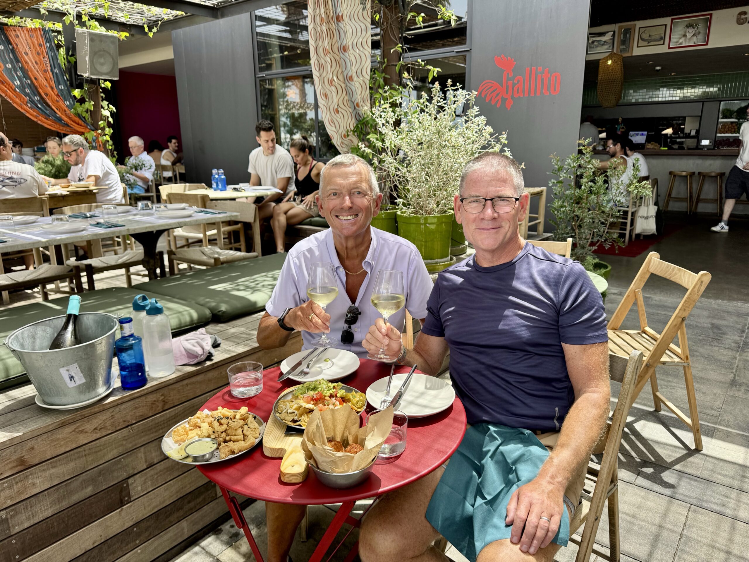 Two men at an outdoor restaurant toast with wine glasses, surrounded by tapas and chips on a red table, greenery and a 'Gallito' sign in the background, smiling at the camera.