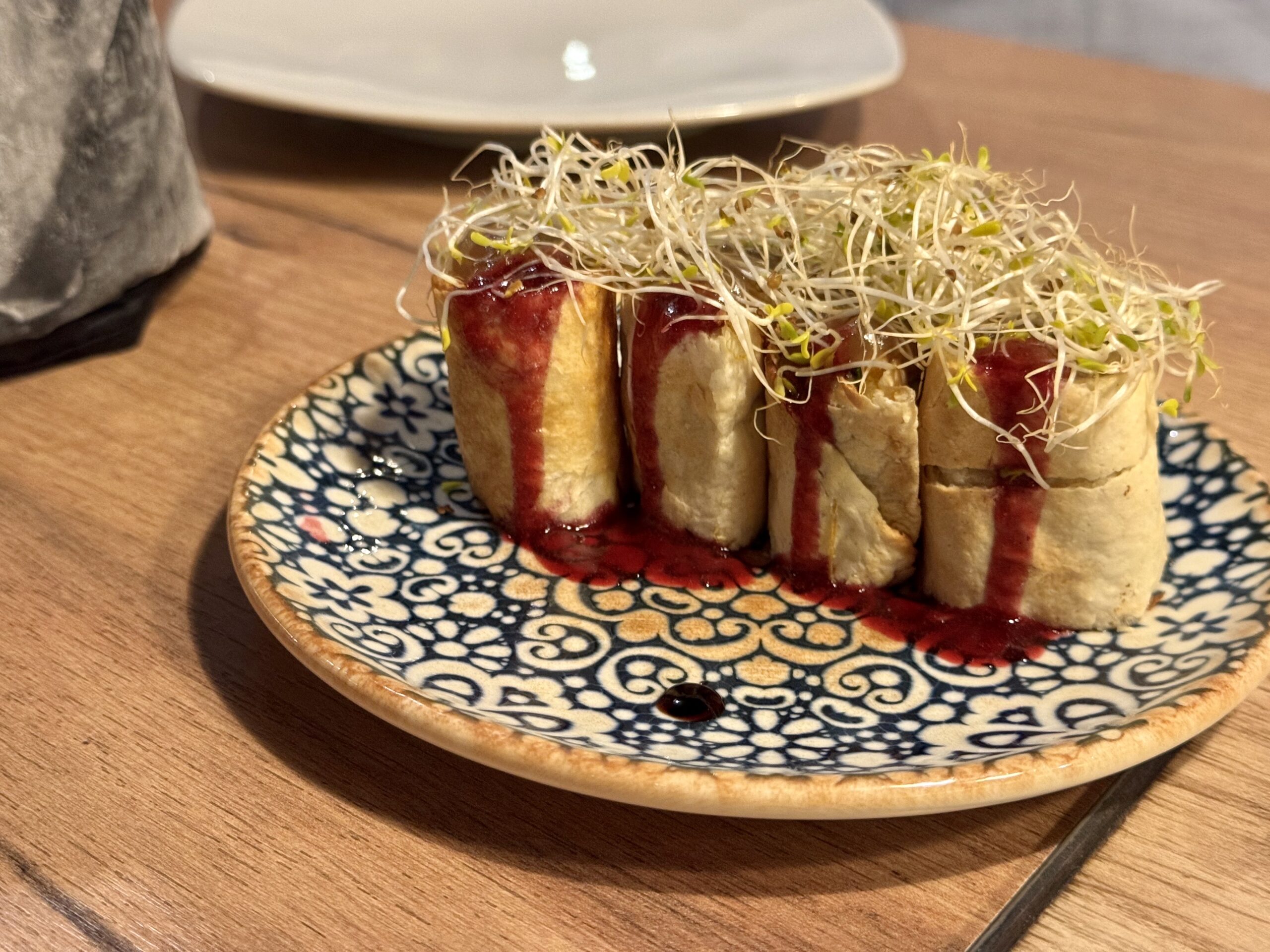 Plate of mini pastry rolls topped with sprouts and a red sauce on a blue patterned plate.