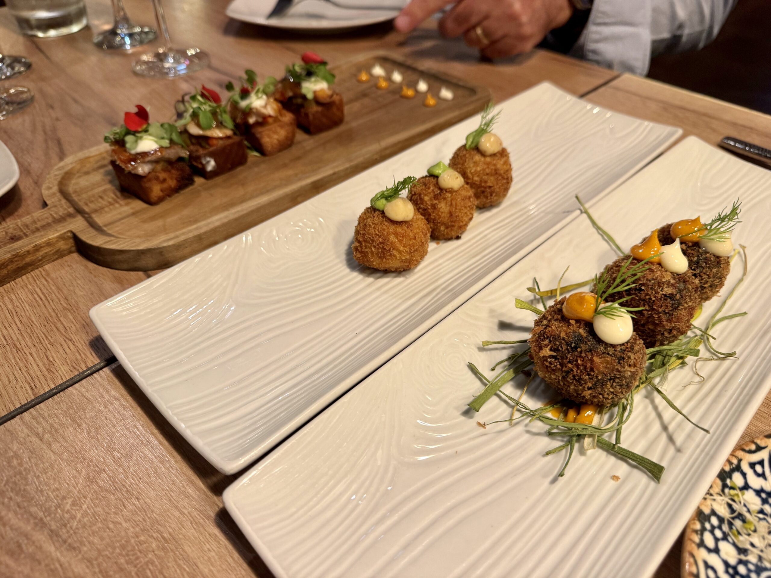 Crispy breaded croquettes with pale dollops and dill on a white rectangular plate; other tapas visible in the background on a wooden board.