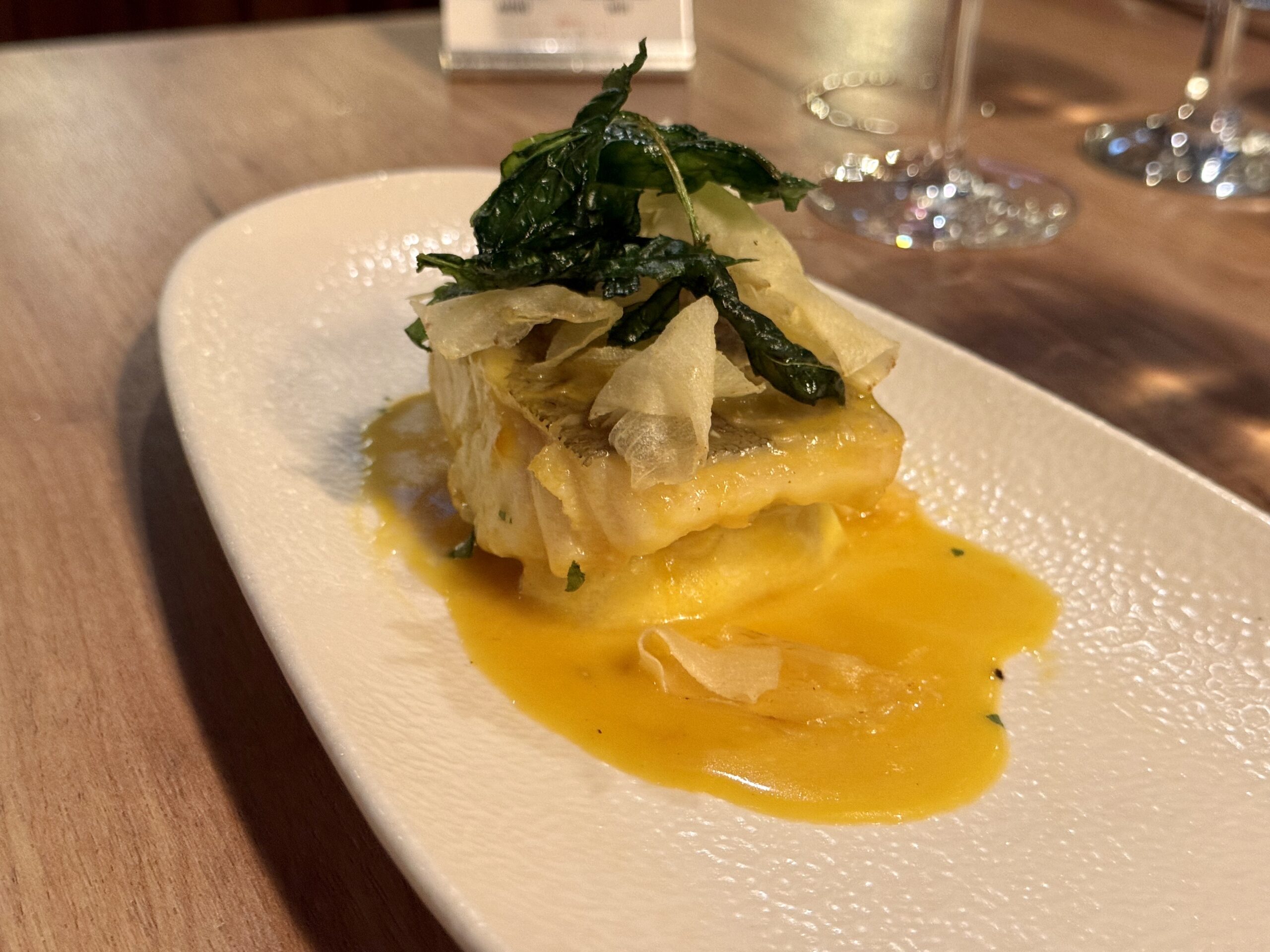 Stack of flaky pastry or fish topped with wilted greens on a bright yellow sauce on a white plate.