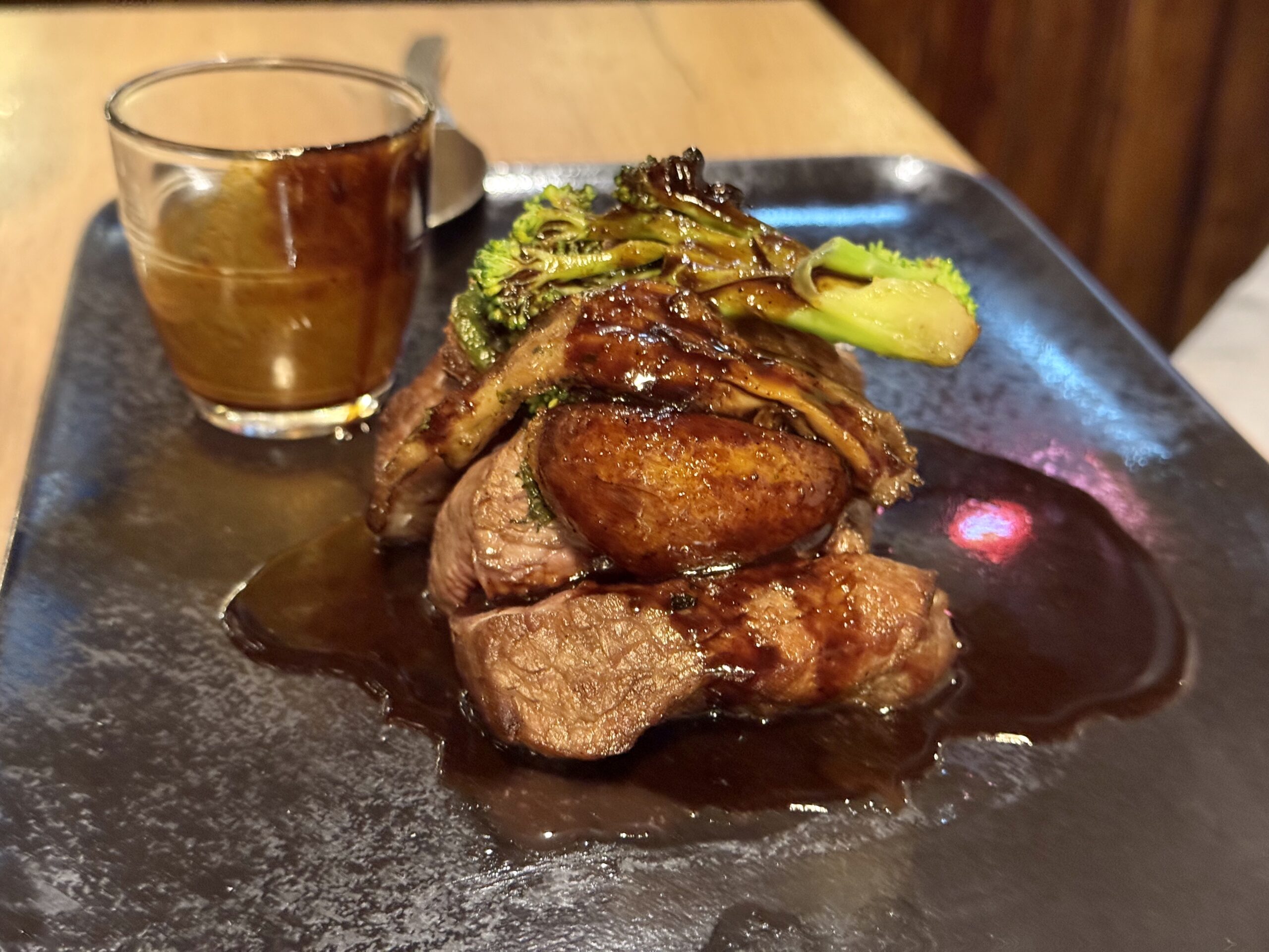 Grilled steak with sautéed greens and glossy sauce on a dark plate, beside a glass of iced drink.