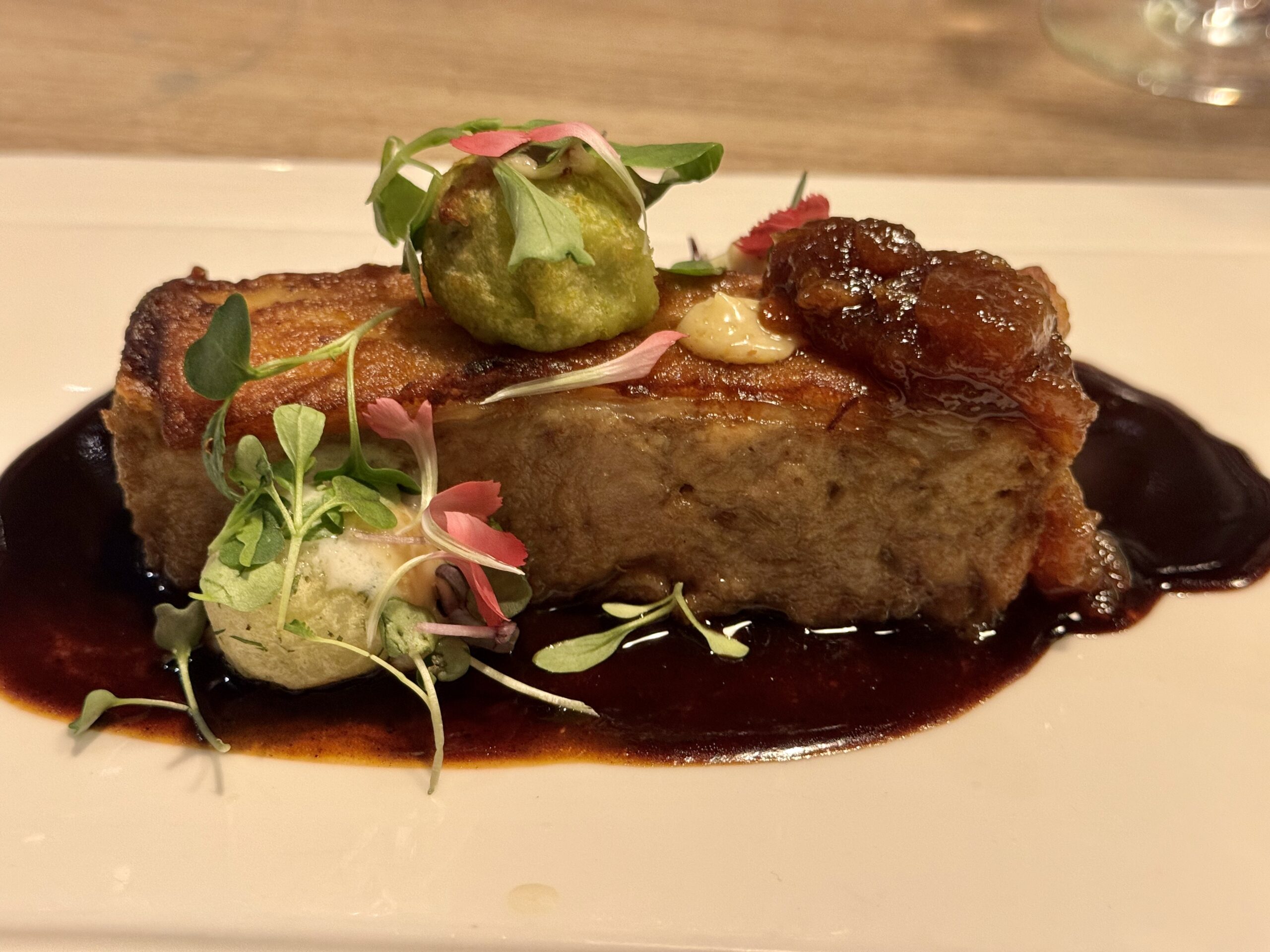 Plate with a seared meat portion, dark glaze, topped by a green fried croquette and microgreens, pink edible petals, and a dollop of pale sauce on the side.