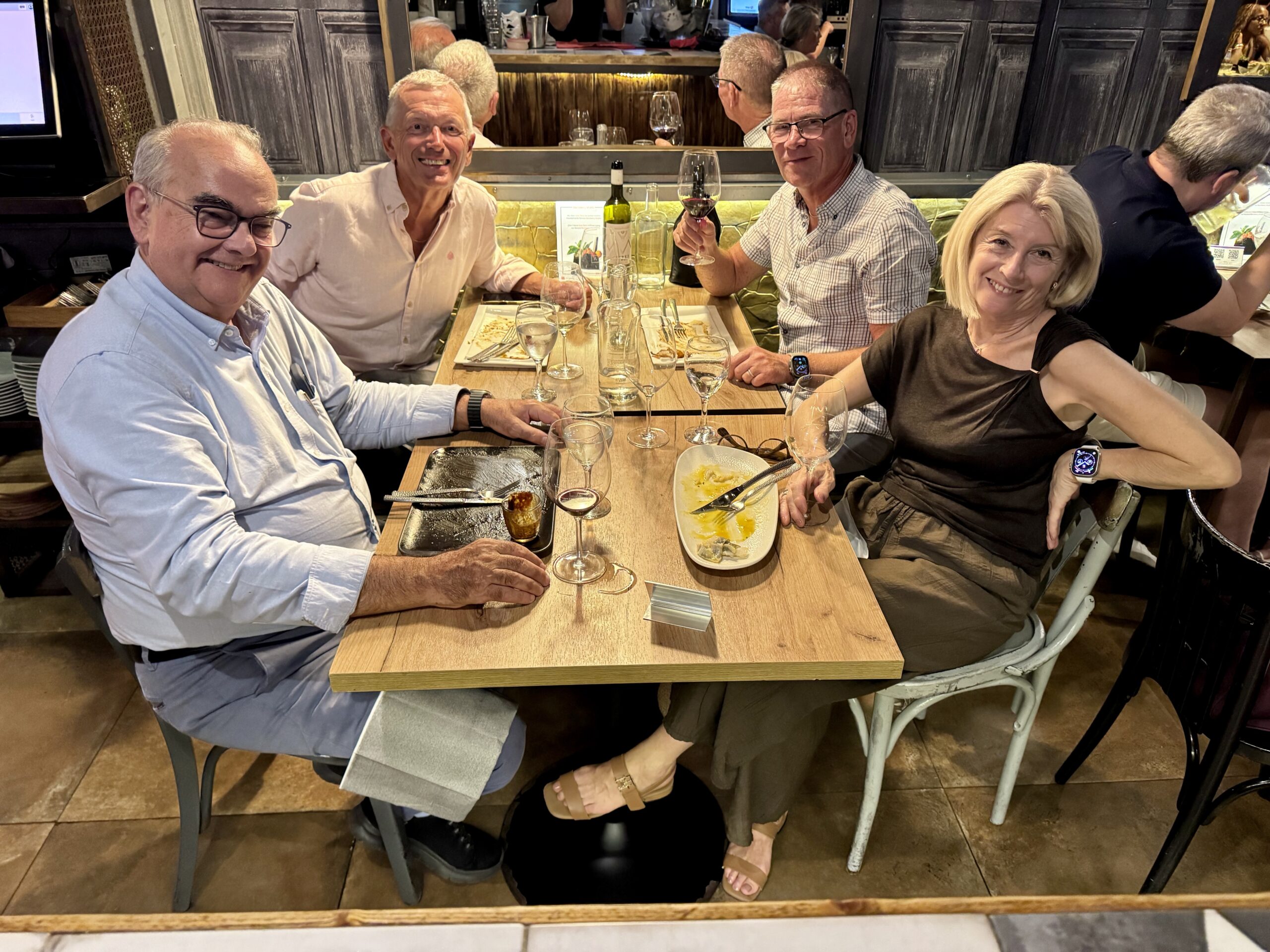 Five adults at a restaurant table smiling for a group photo; wine glasses, plates, and a bottle on the table.