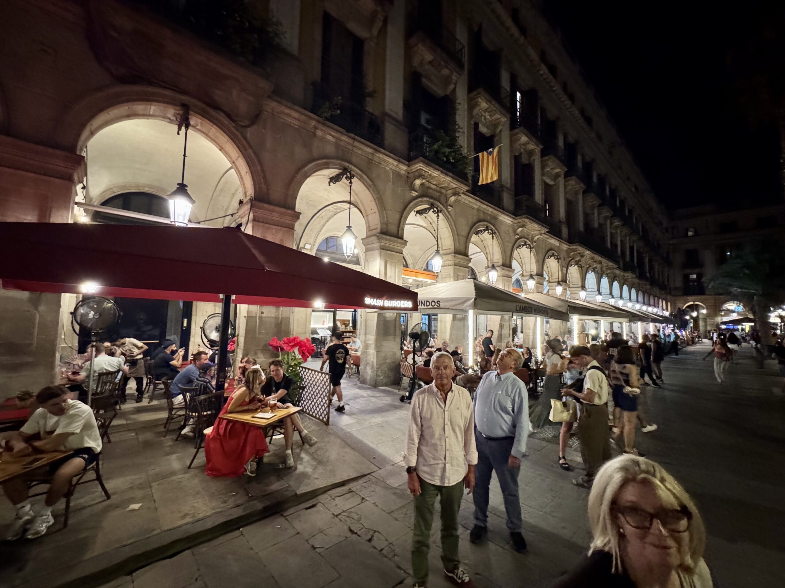 Night street scene of a crowded outdoor cafe under stone arches with warm lantern light.