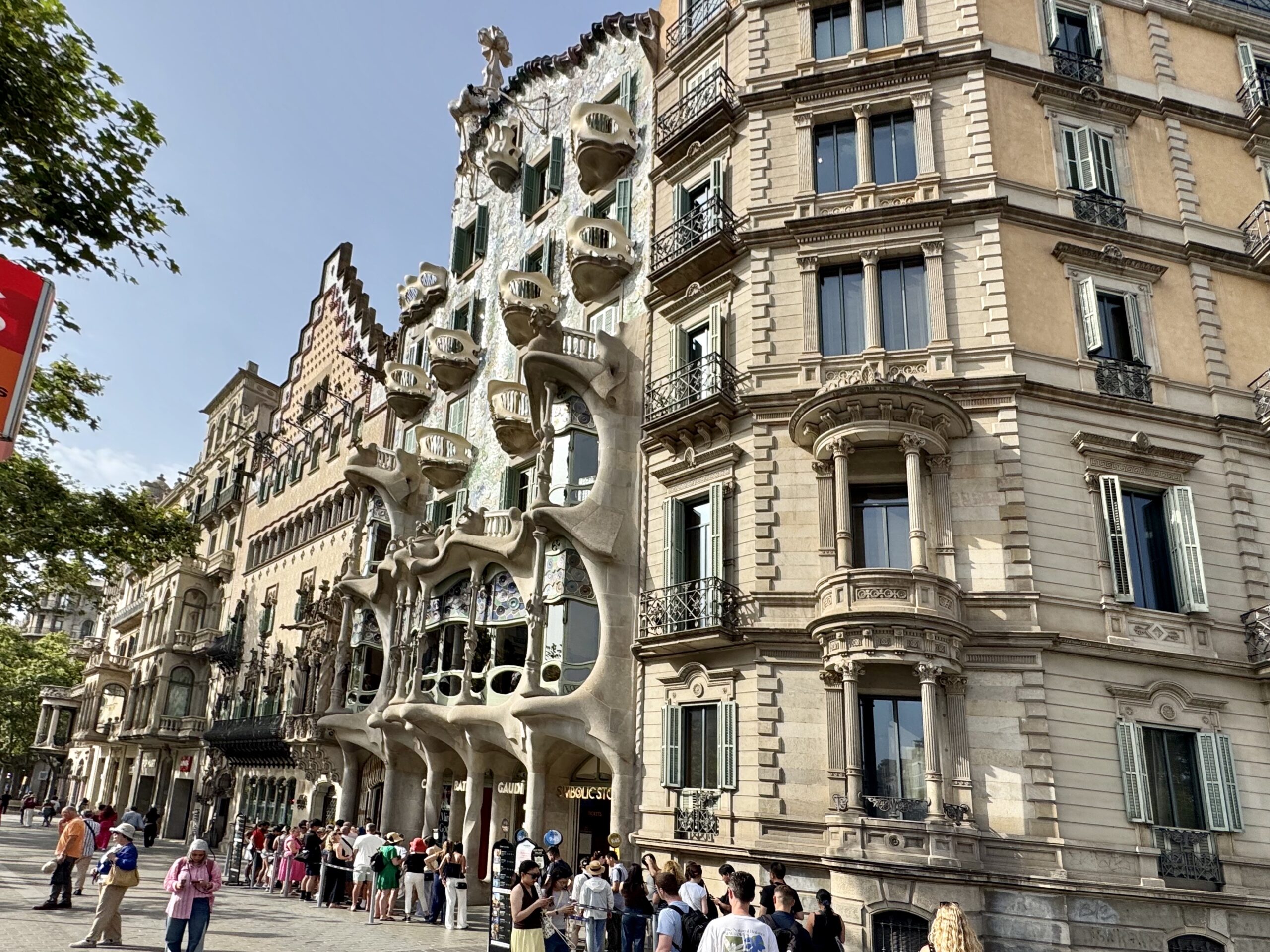 Front facade of Gaudí's Casa Batlló on Passeig de Gràcia with sculpted balconies and wave-like stonework; people queue along the sidewalk.