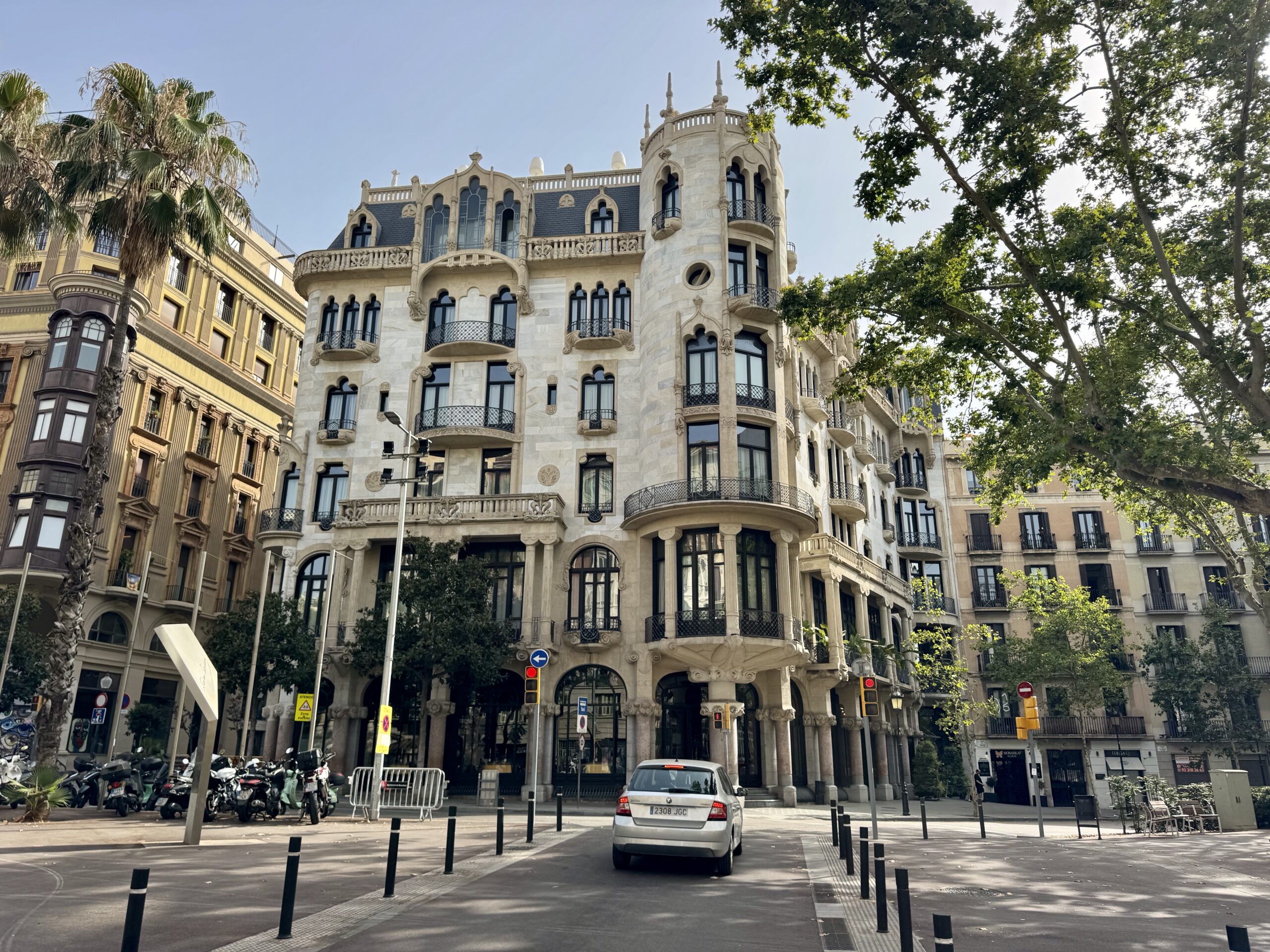 Ornate beige stone building with rounded turret and decorative balconies on a sunny street, lined with palm trees and cars.