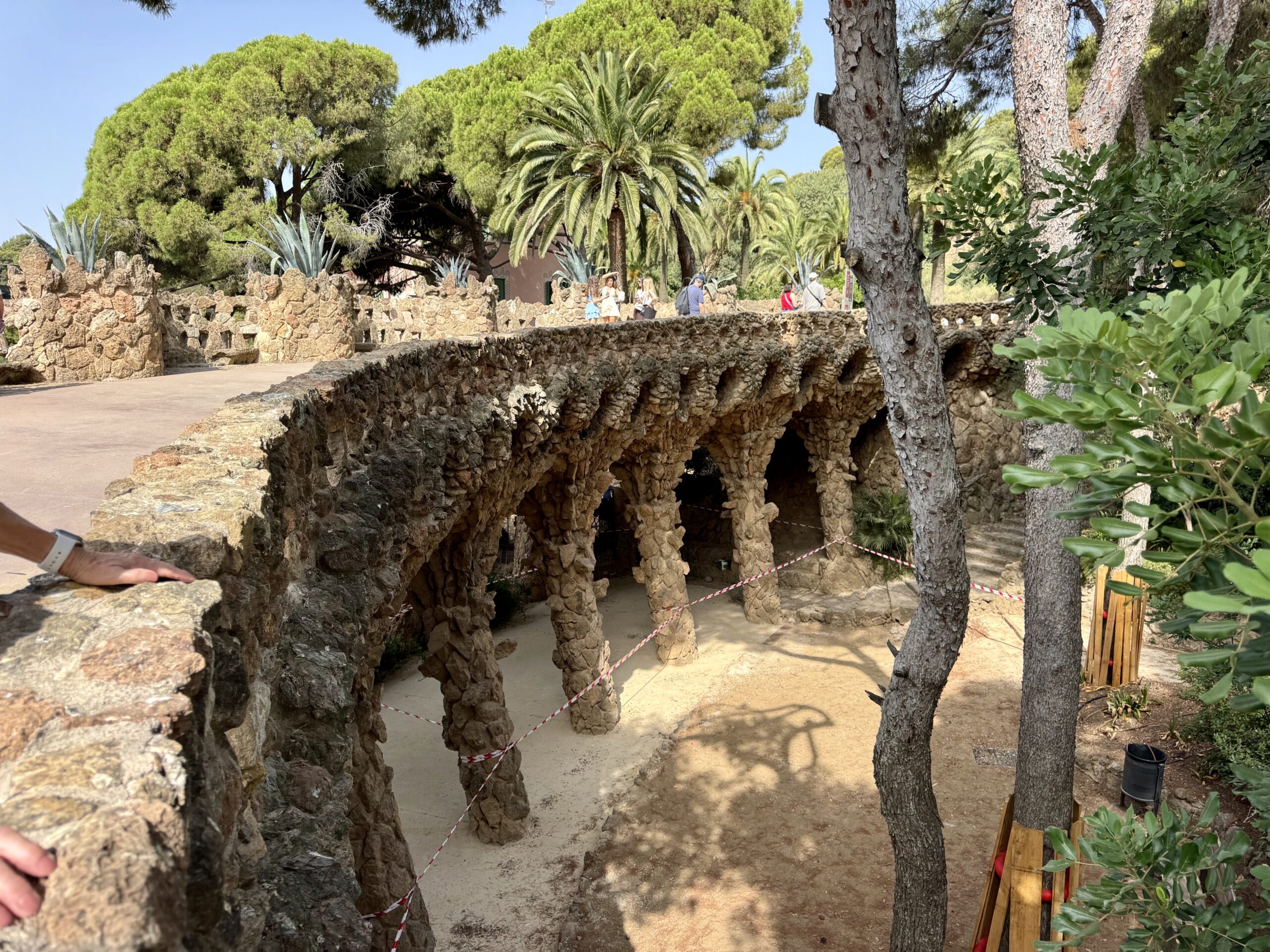 Stone stone-arch walkway with carved columns, green palm trees, and visitors in the background on a sunny day.
