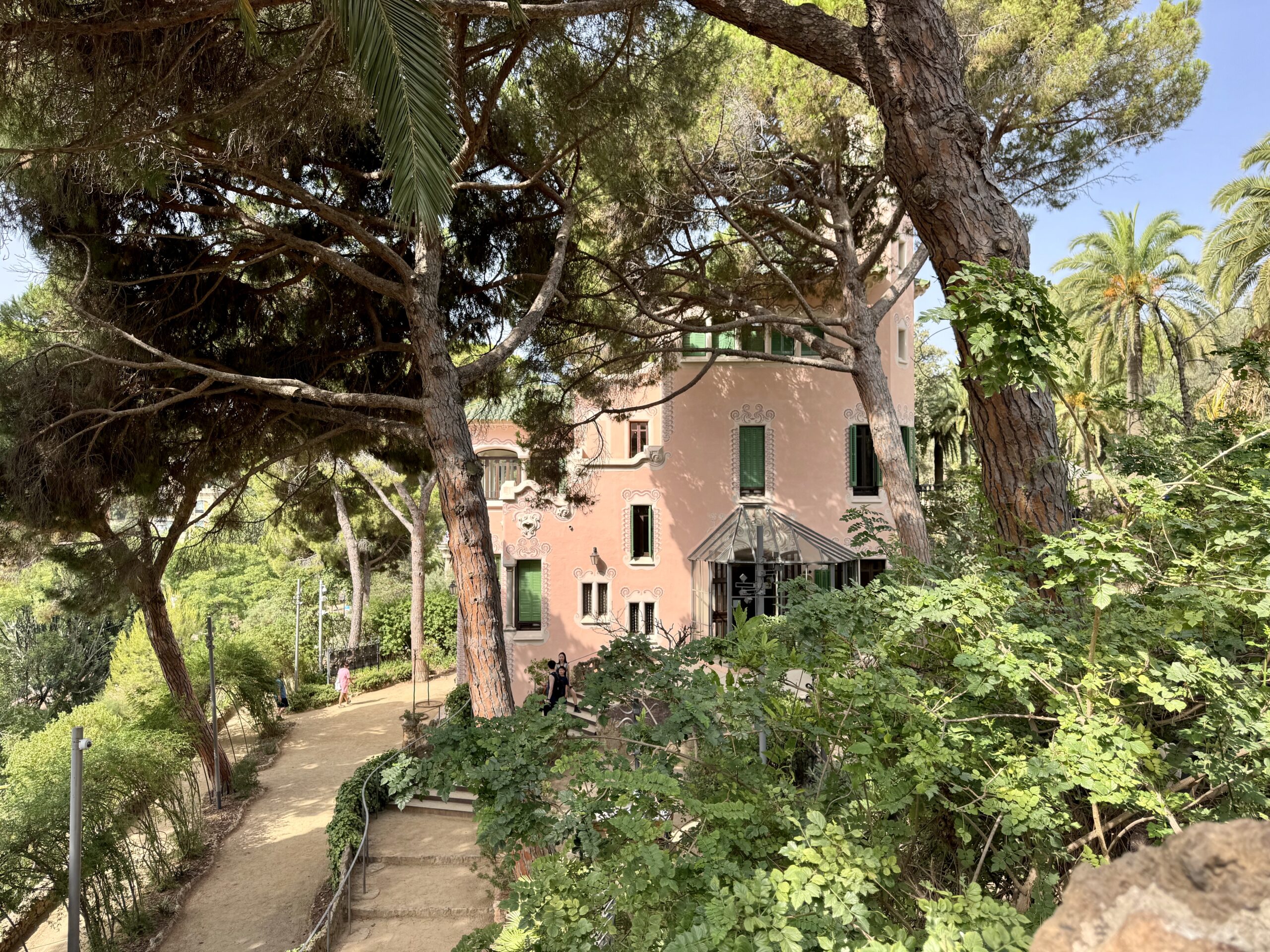 Pink stucco house with green shutters partially visible through dense trees and a sunlit garden path in front.