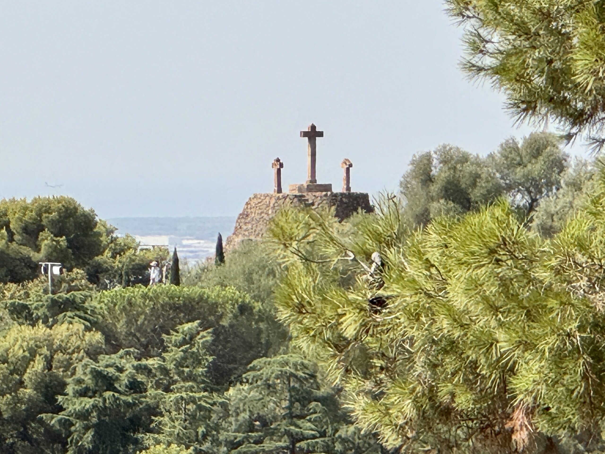 Stone hilltop monument featuring a central cross, with two smaller pillars, surrounded by trees and greenery.