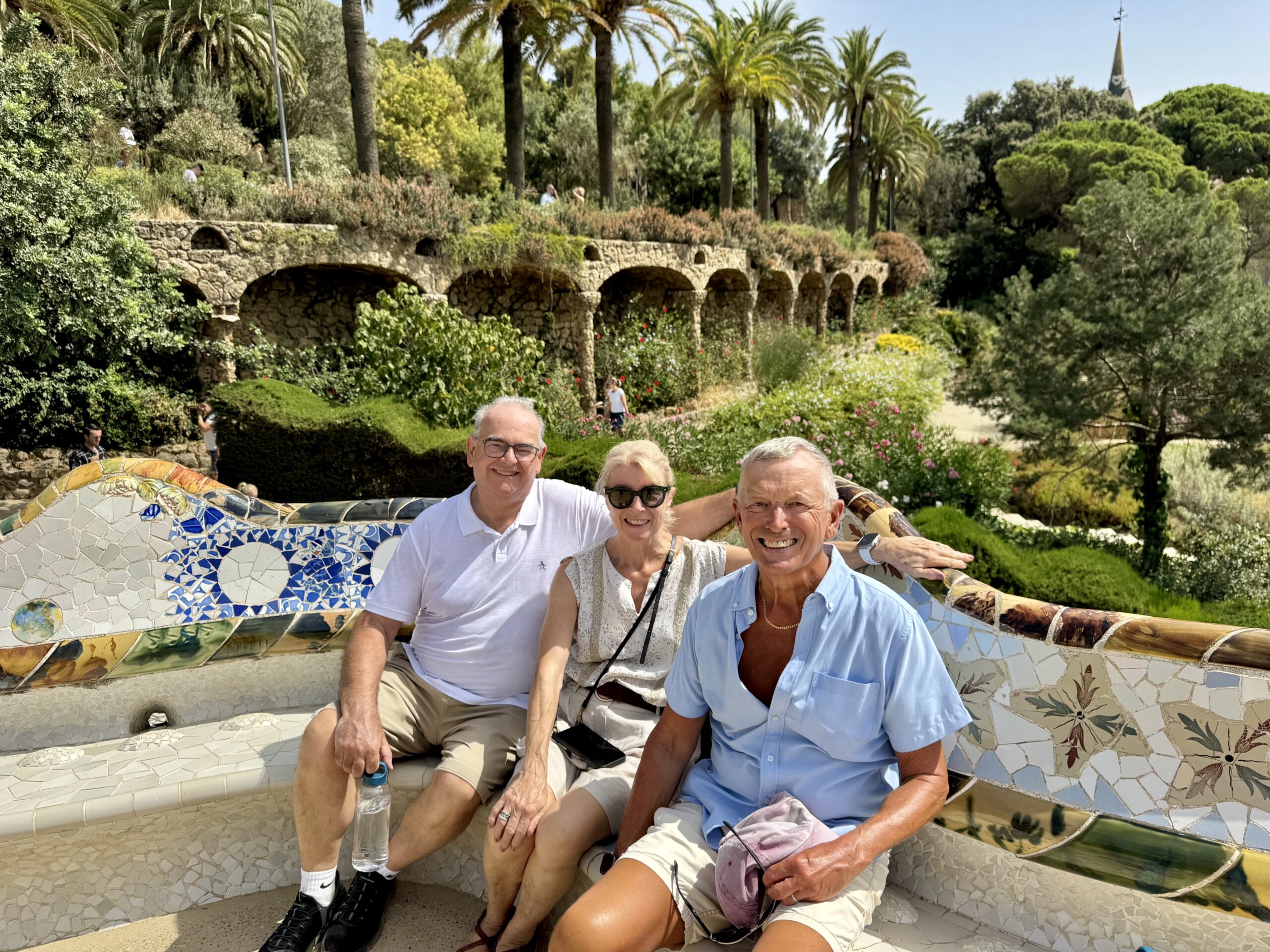 Four adults smiling on a colorful mosaic bench in a sunny garden with stone arches and palm trees in the background.