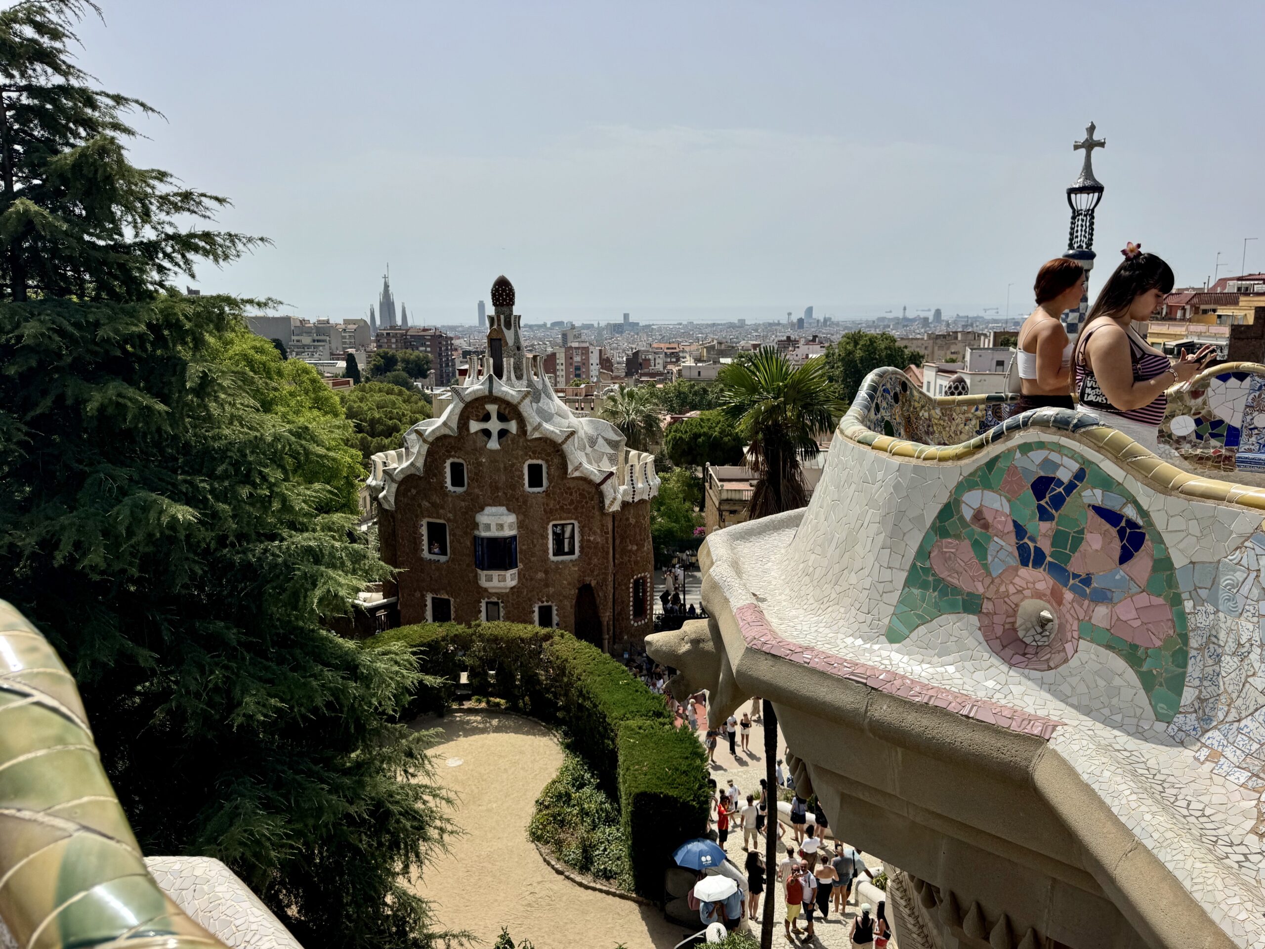 View over a colorful mosaic serpent bench at Park Güell with visitors and Barcelona skyline in the distance under a clear sky.