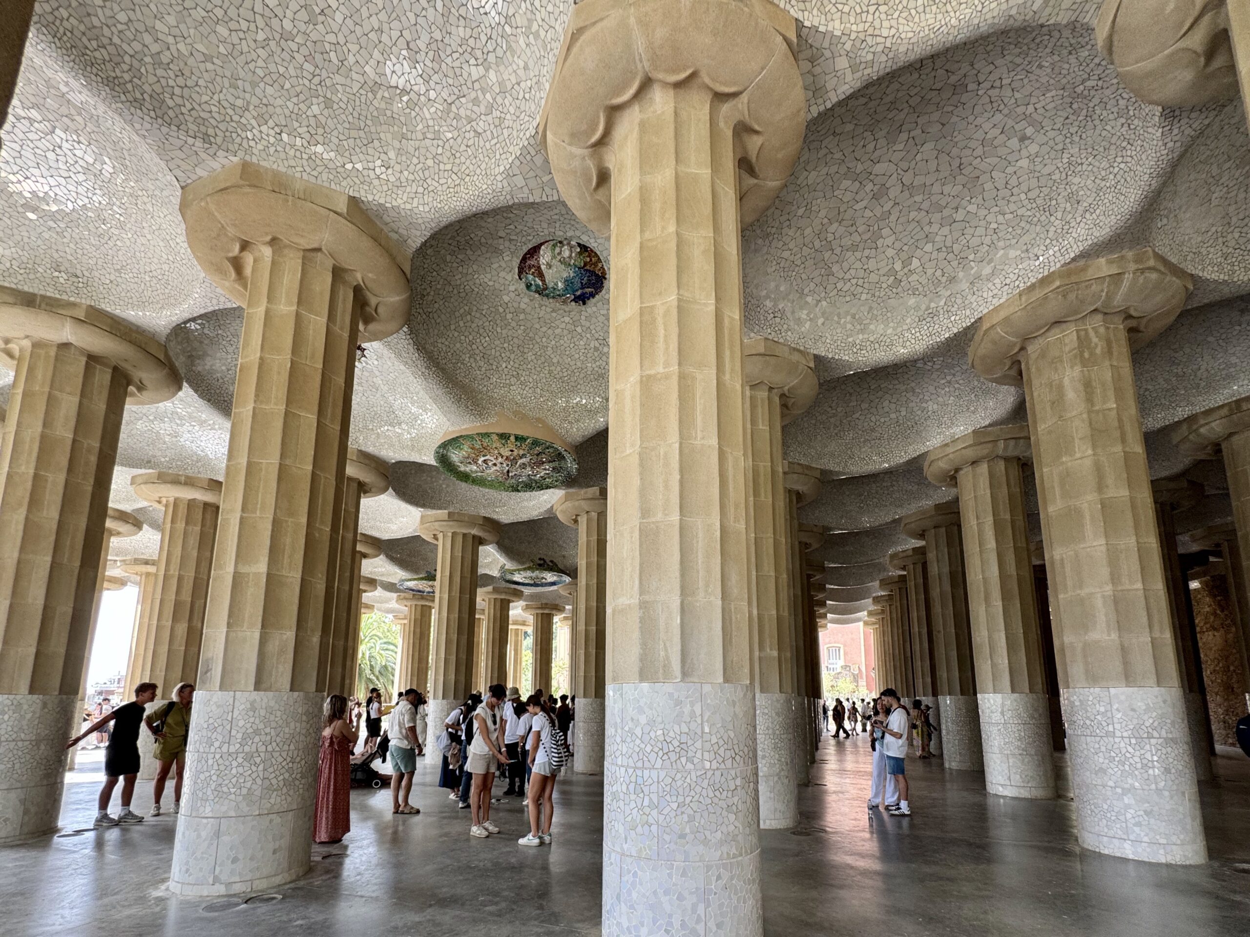 Interior of Sagrada Família hypostyle hall with dozens of stone columns and mosaic-covered ceilings, and visitors walking beneath them.