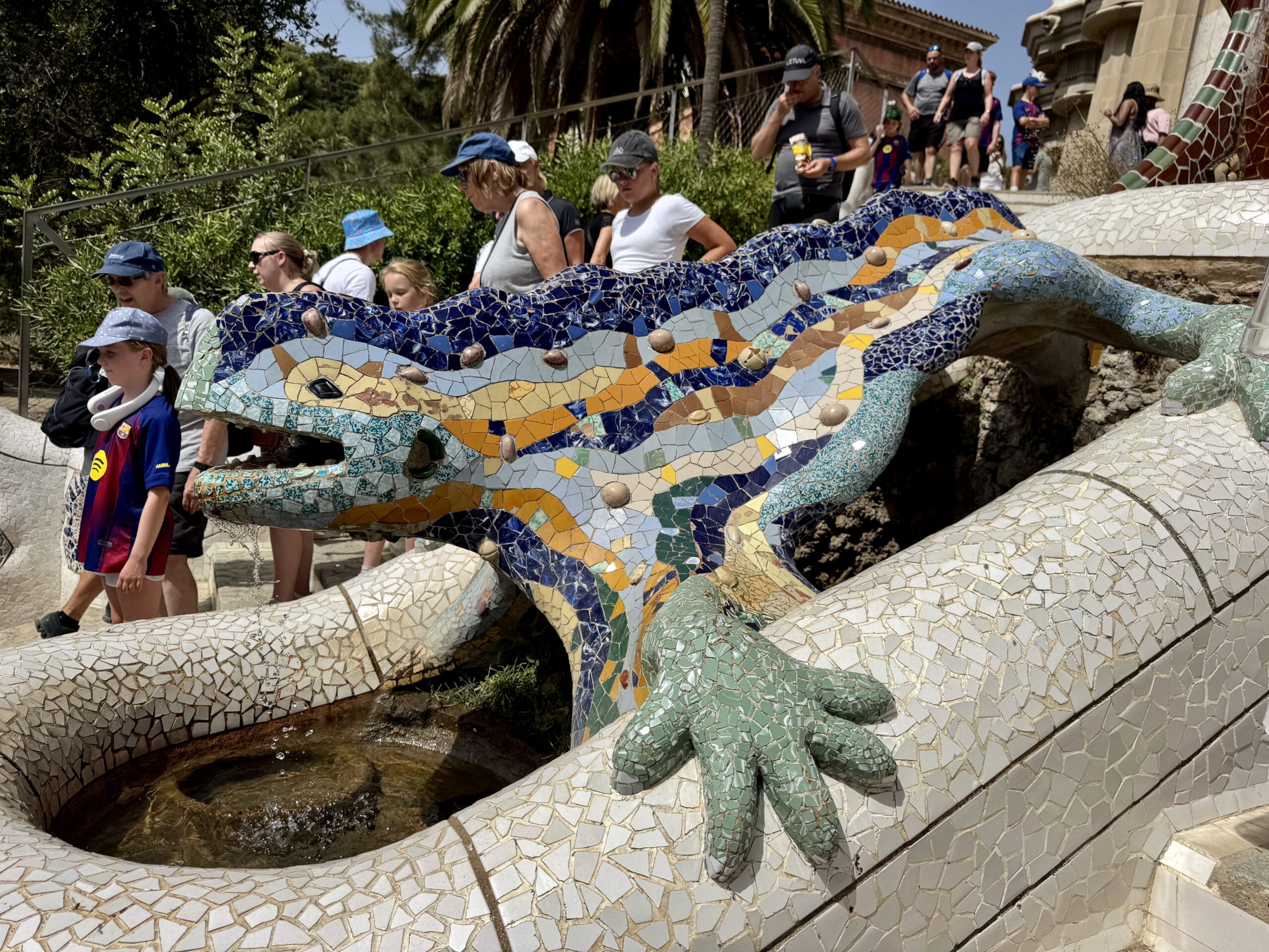 Colorful mosaic dragon sculpture with people standing on its back in a sunny outdoor setting, a park area in the background.