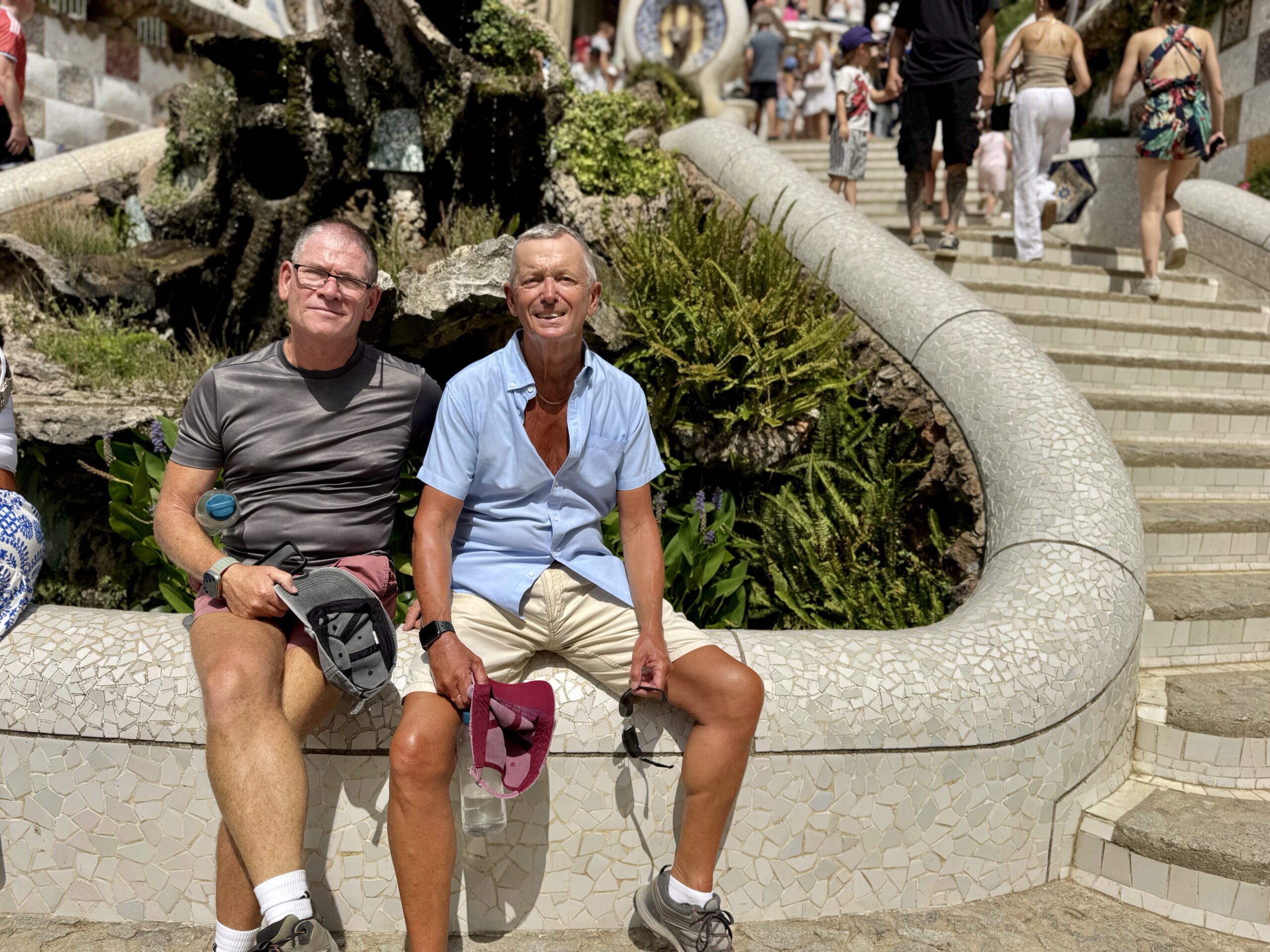 Two older men sit on a curved mosaic stone bench in a sunny outdoor area, with people walking up steps behind them.
