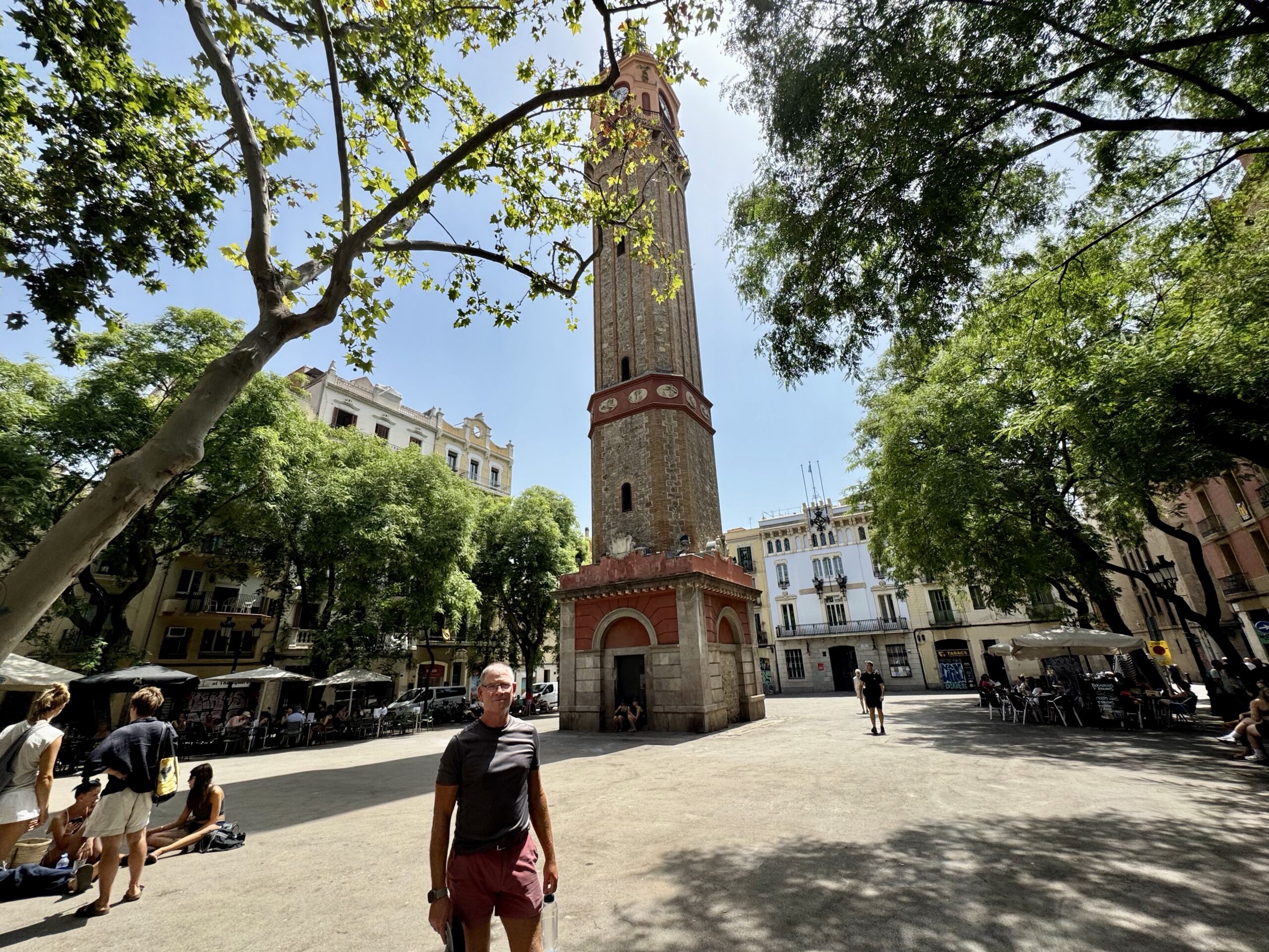 Clock tower rising above a sunny plaza framed by leafy trees and outdoor cafe seating with people nearby, urban scene in daytime.
