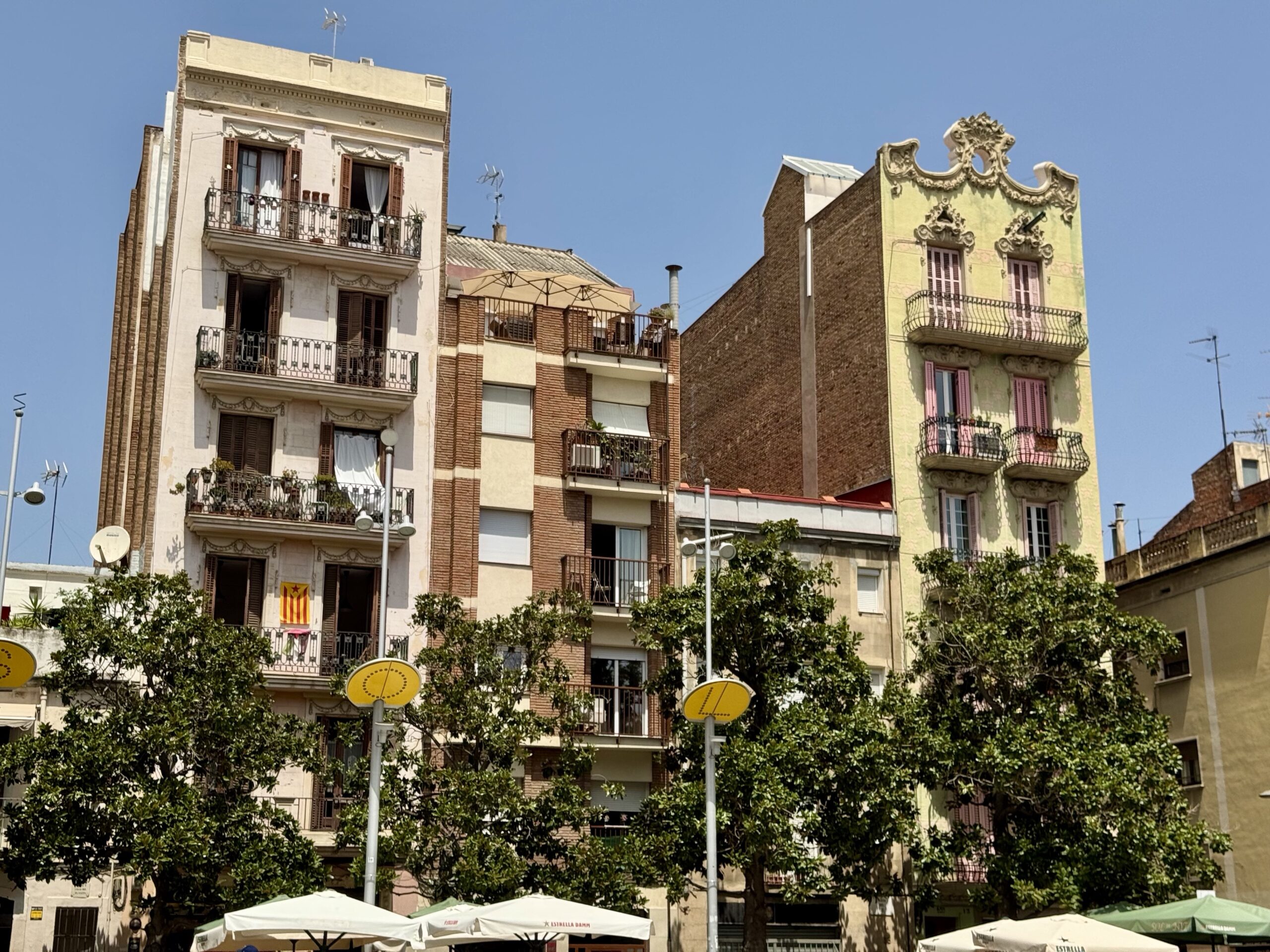 Facade of a row of ornate European buildings with balconies, under a clear blue sky; outdoor café umbrellas visible at street level in the foreground.
