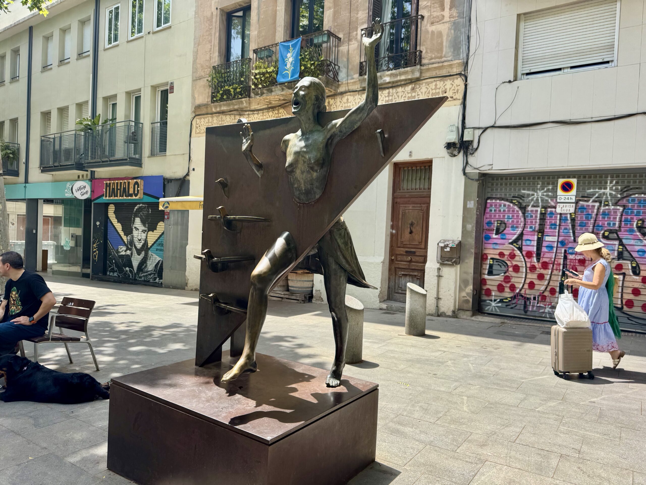 Bronze statue of a dancing figure with one arm raised, mounted on a rust-colored angled panel in a city square with storefronts in the background.