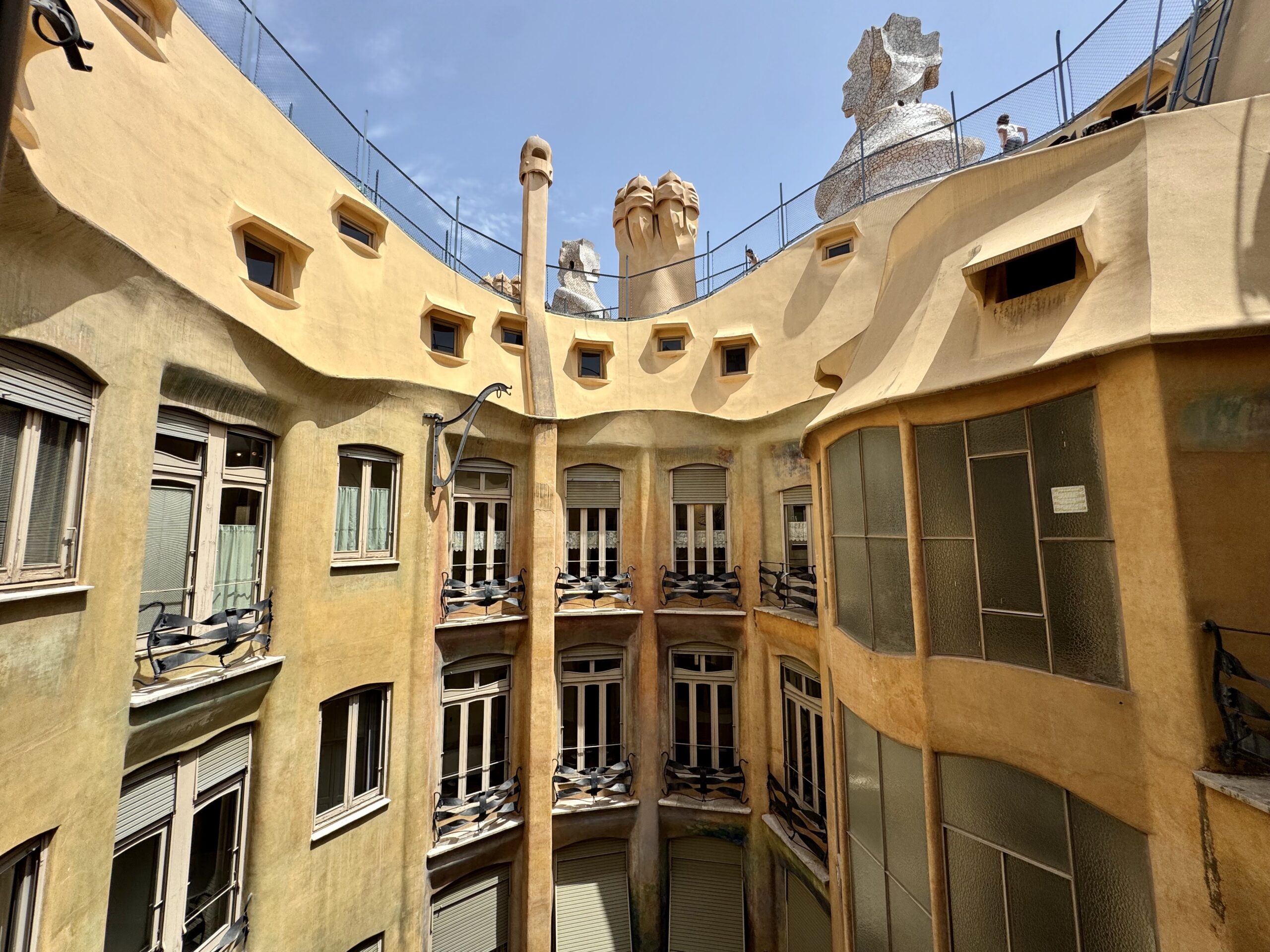 Gaudí-inspired courtyard with curved ochre walls, small square windows, and balconies with metal chairs against a blue sky