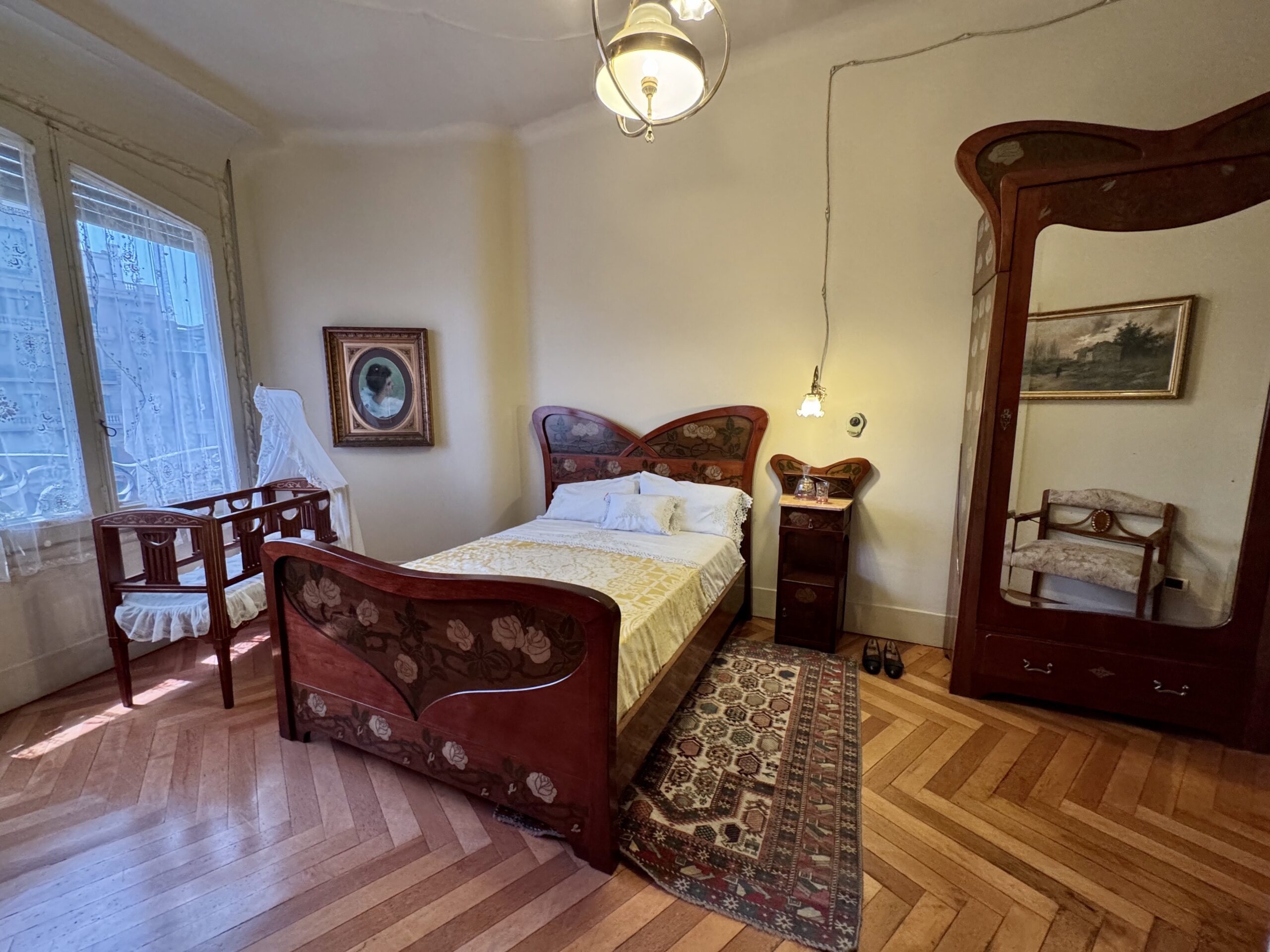 Vintage bedroom with an ornate wooden bed, matching dresser and mirror, and a large window with lace curtains.