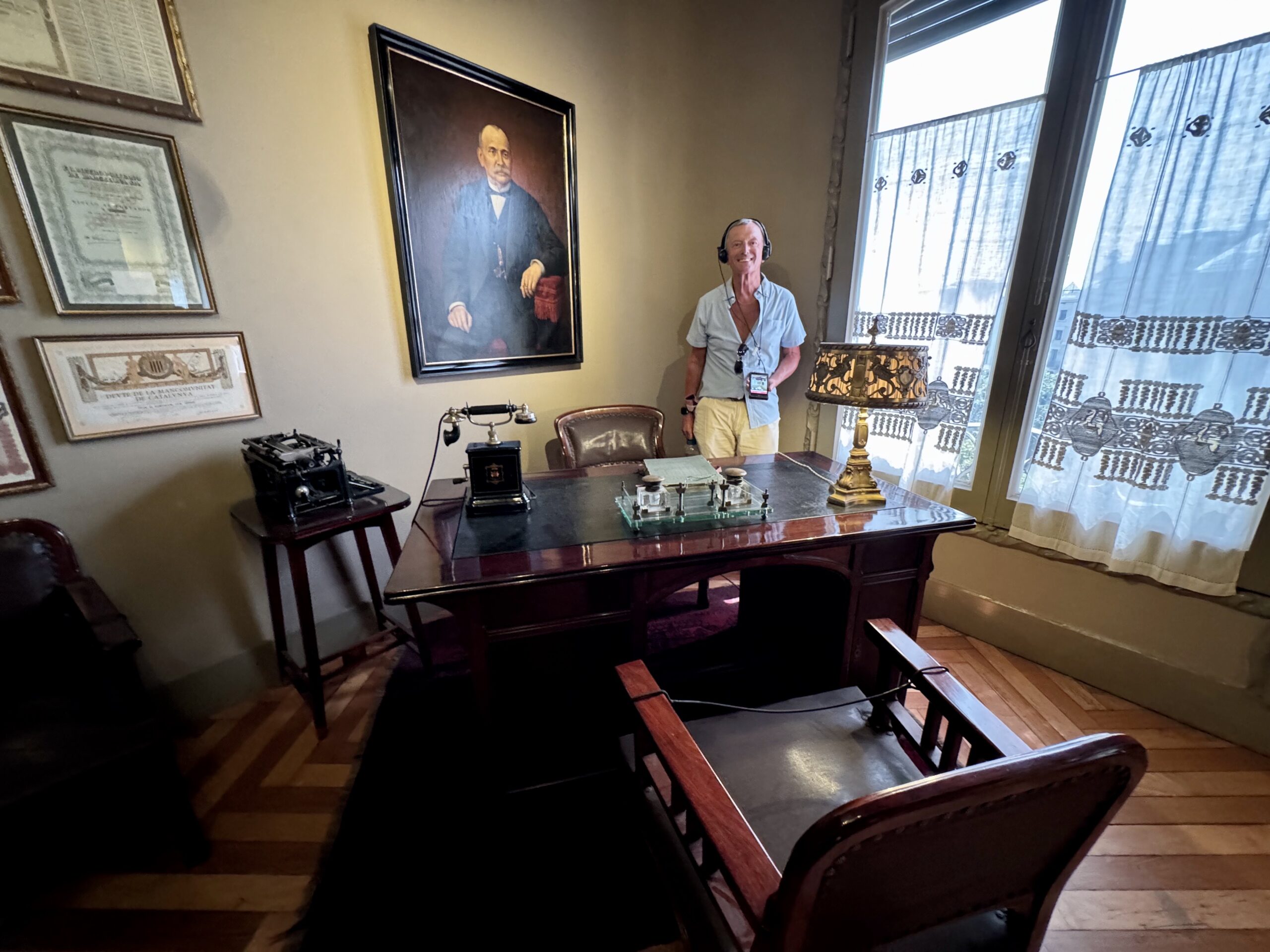 Person wearing a headset stands beside an antique desk in a vintage office with framed portraits on the wall and tall windows with sheer curtains.