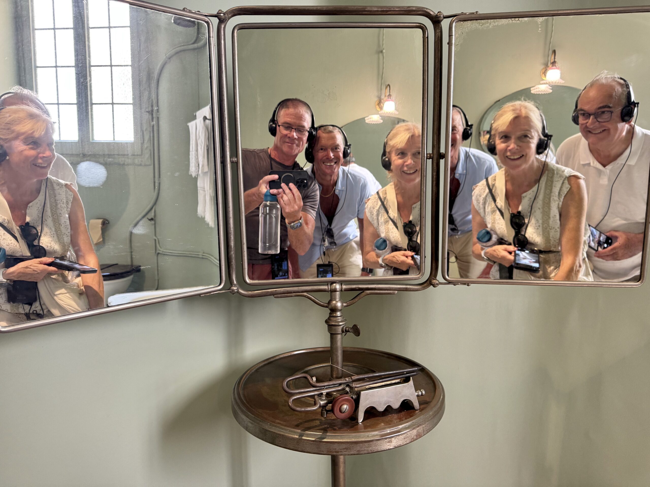 Six adults wearing headphones lean toward a vintage three-panel mirror, posing for a group photo in a pastel-green bathroom.
