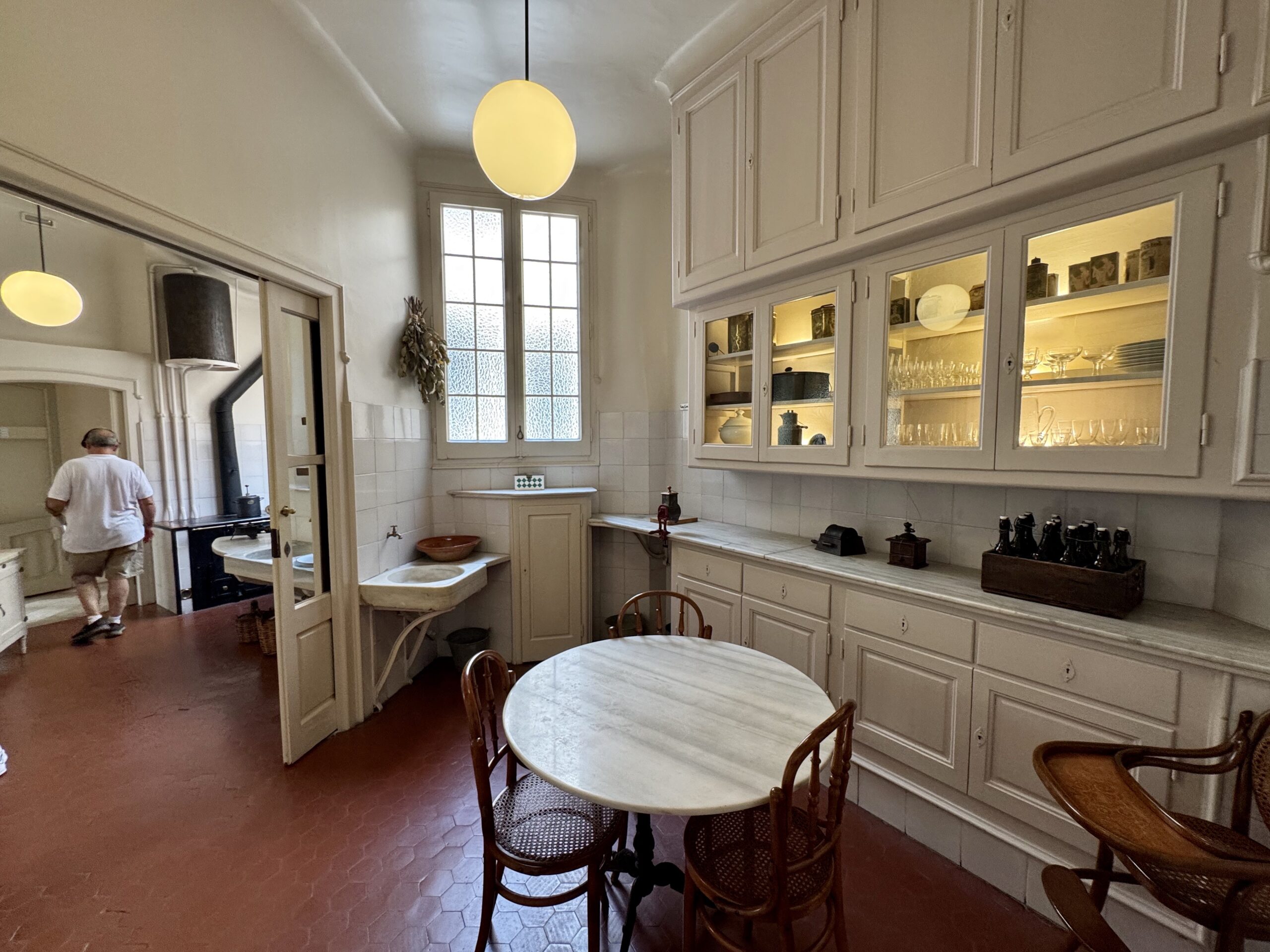 Vintage kitchen with glass-front cabinets, round marble tables, and a man walking away under a warm pendant light.