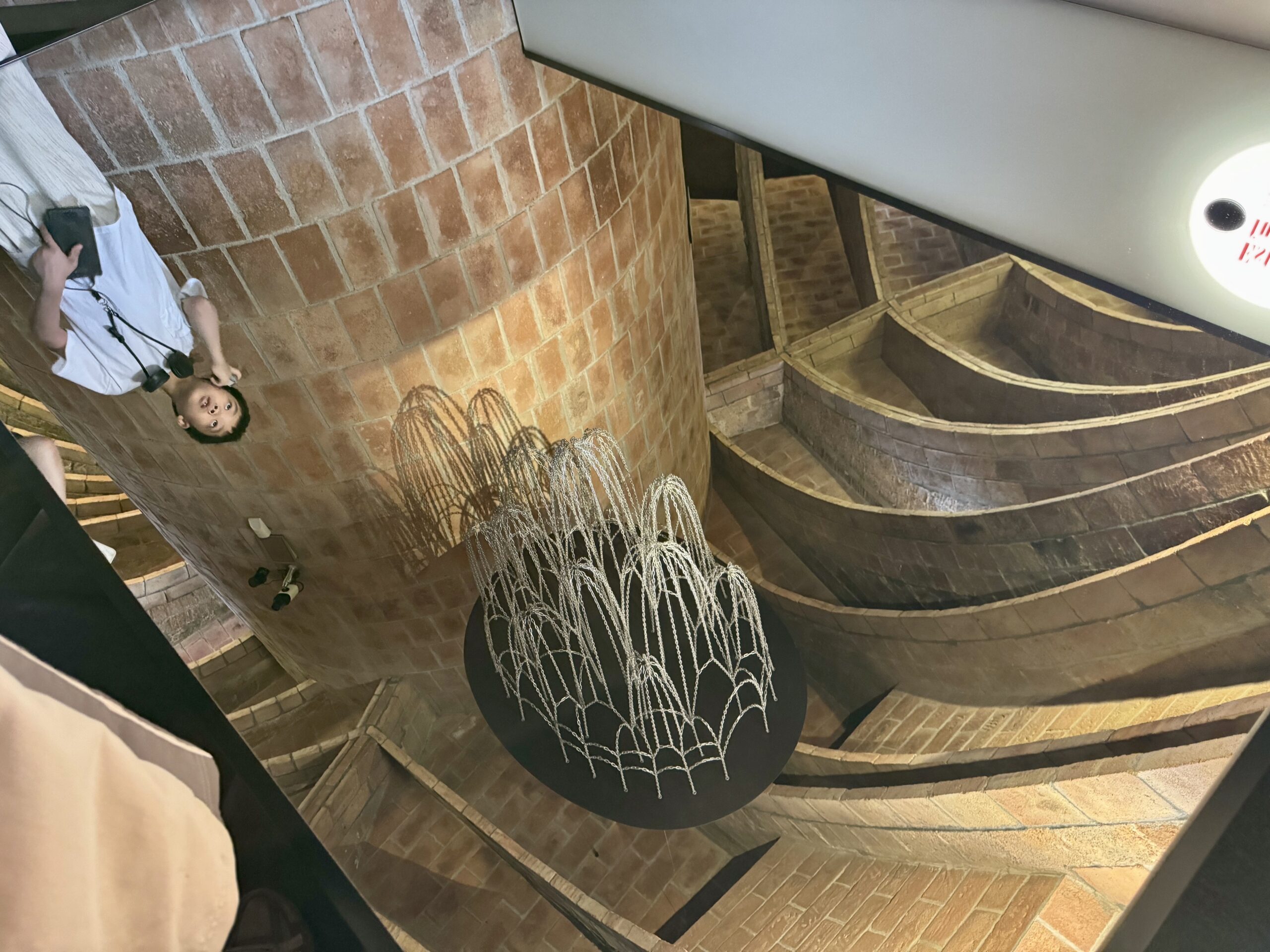 Person in a white shirt stands on a curved brick stairwell, holding a phone, with a decorative wire sculpture in the foreground.