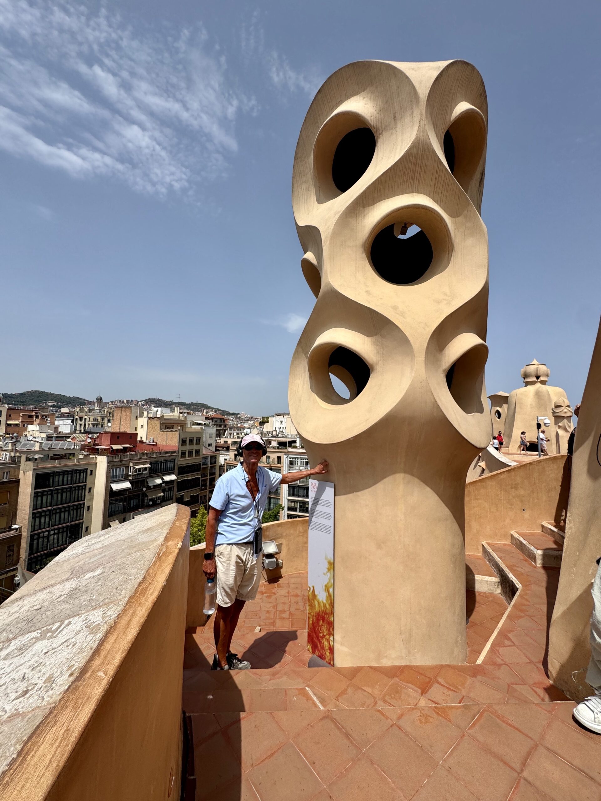 Tourist in a light blue shirt and hat beside a tall beige sculpture with circular hollow openings on a rooftop walkway, city below under a blue sky.