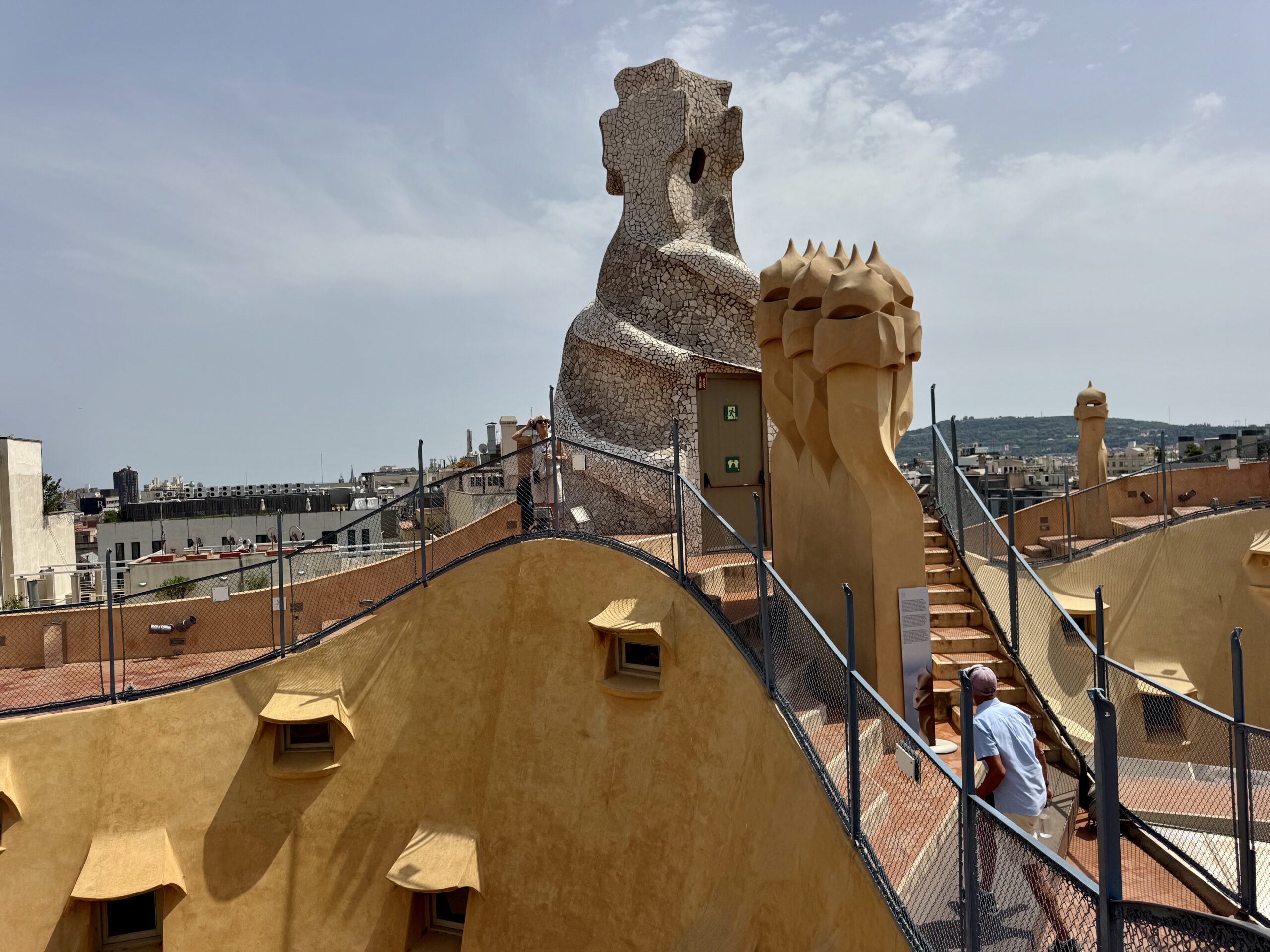 Rooftop terrace with a tall cracked-mosaic sculpture and beige sculpted pillars, fenced walkways, and city buildings in the background.