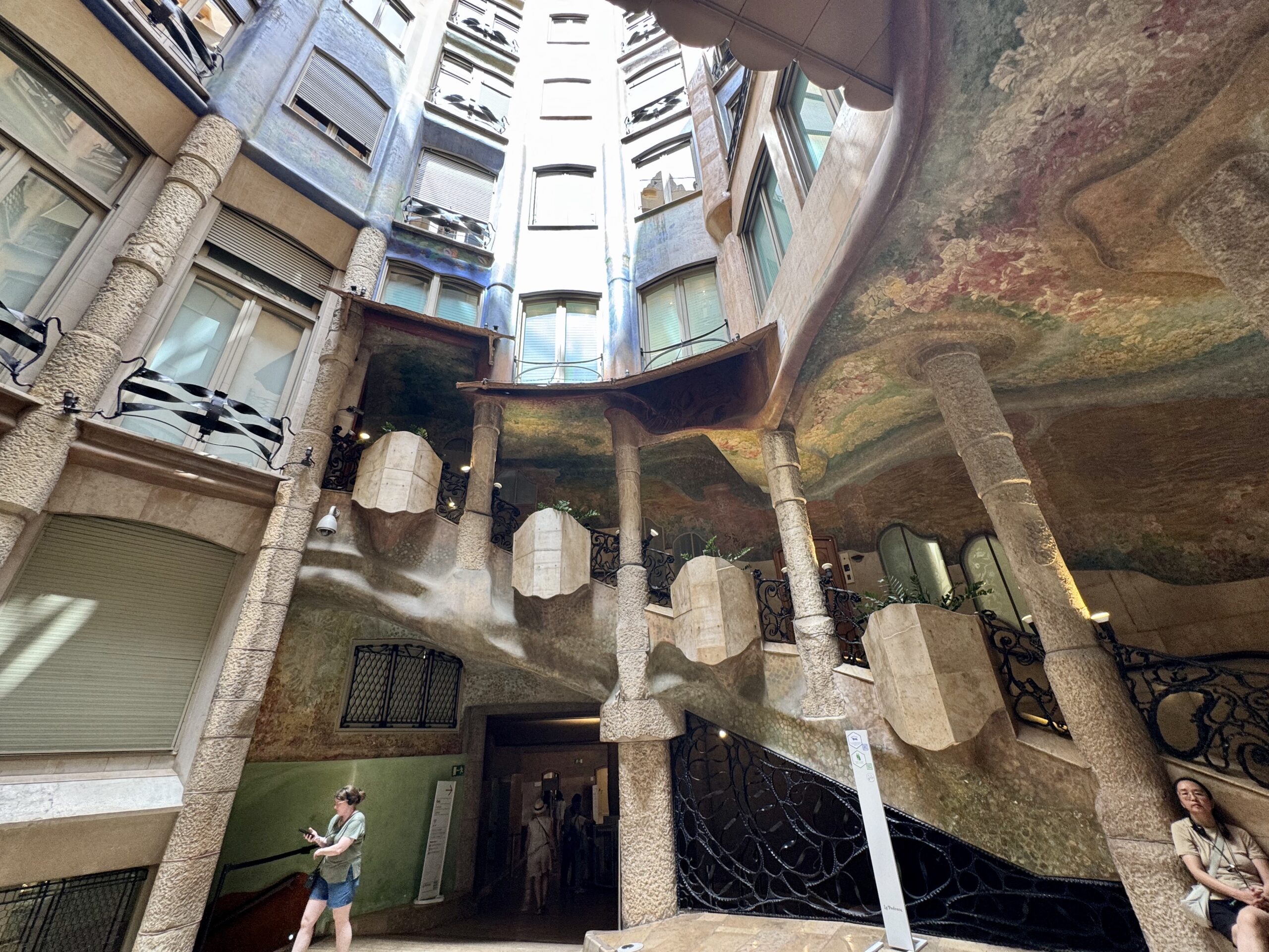 Indoor courtyard with tall textured stone columns, curved balconies, and pastel mural walls seen from below, with visitors nearby.