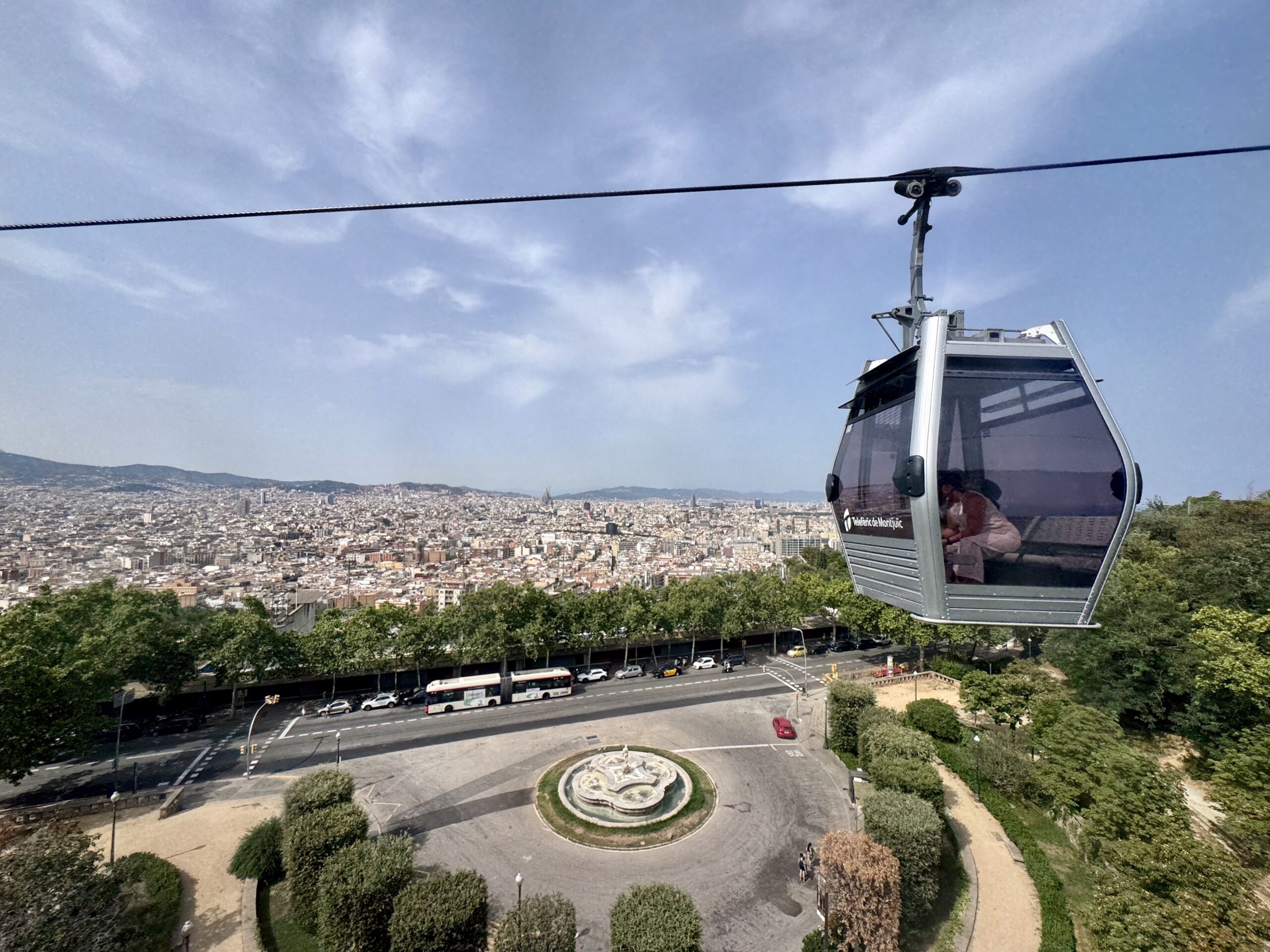 Cable car gondola flying above a circular roundabout with green trees and a city skyline in the distance.