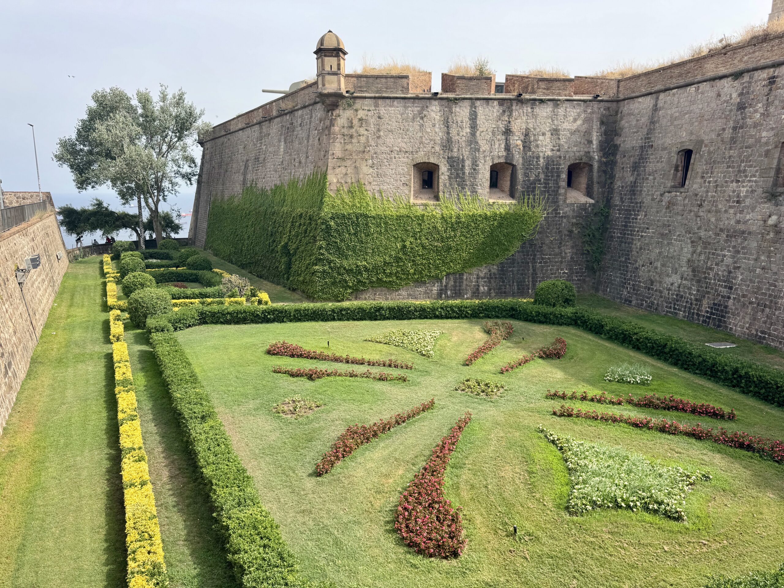Stone fortress wall with ivy-covered base, and formal garden beds with trimmed hedges and green lawn in front.
