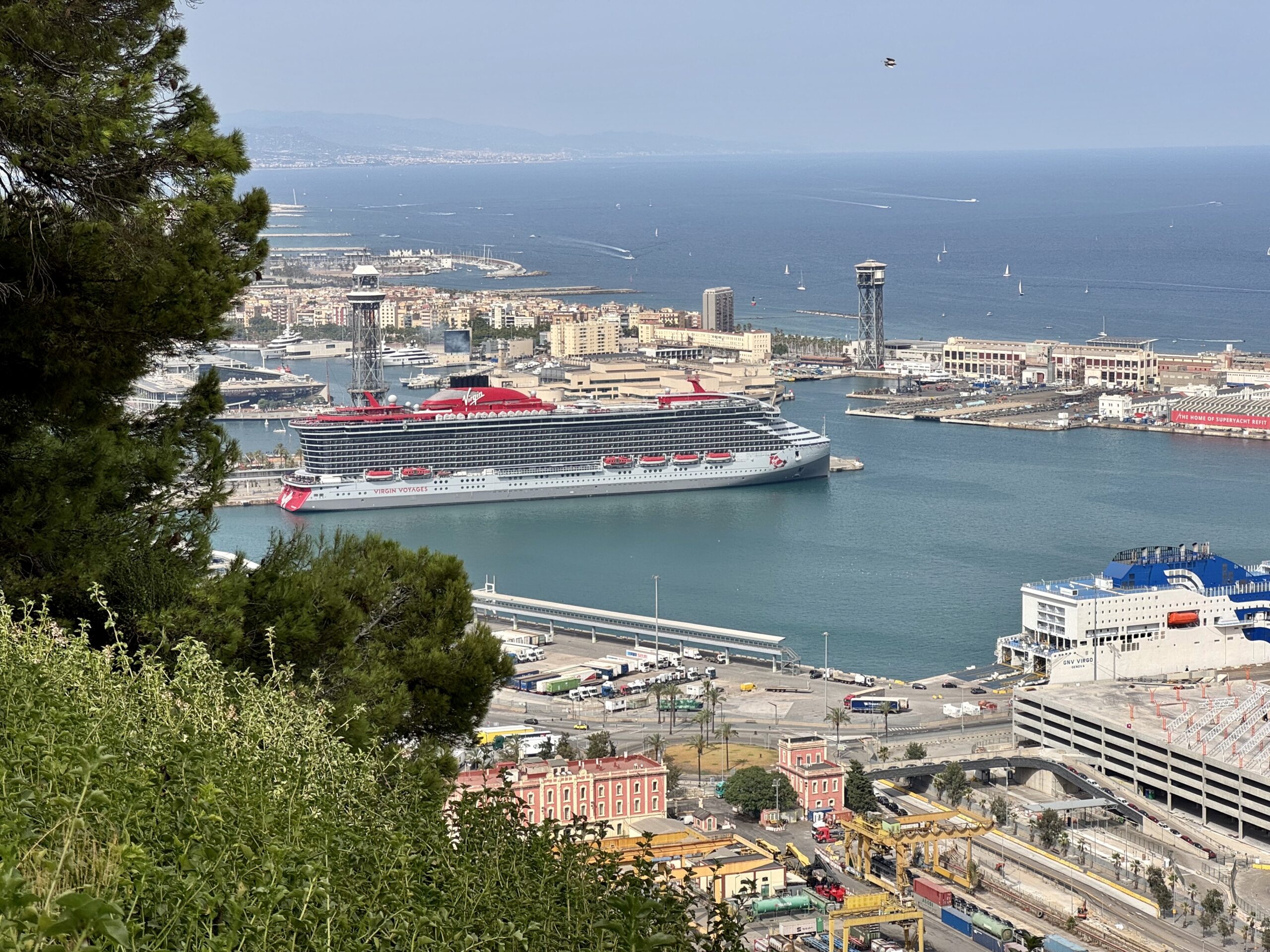A large Virgin Voyages cruise ship docked at a busy harbor with city buildings, cranes, and distant hills, seen from a green hillside foreground.
