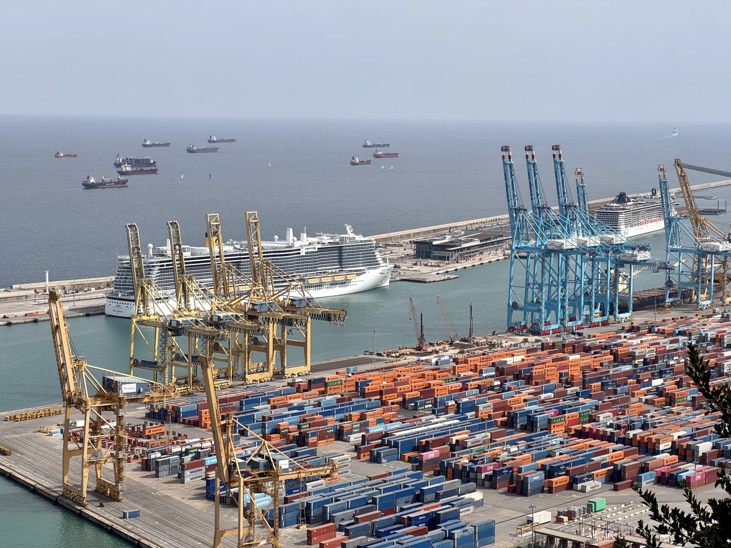 A busy port with tall blue cranes, rows of colorful shipping containers, and several ships in the water near a docked cruise liner.