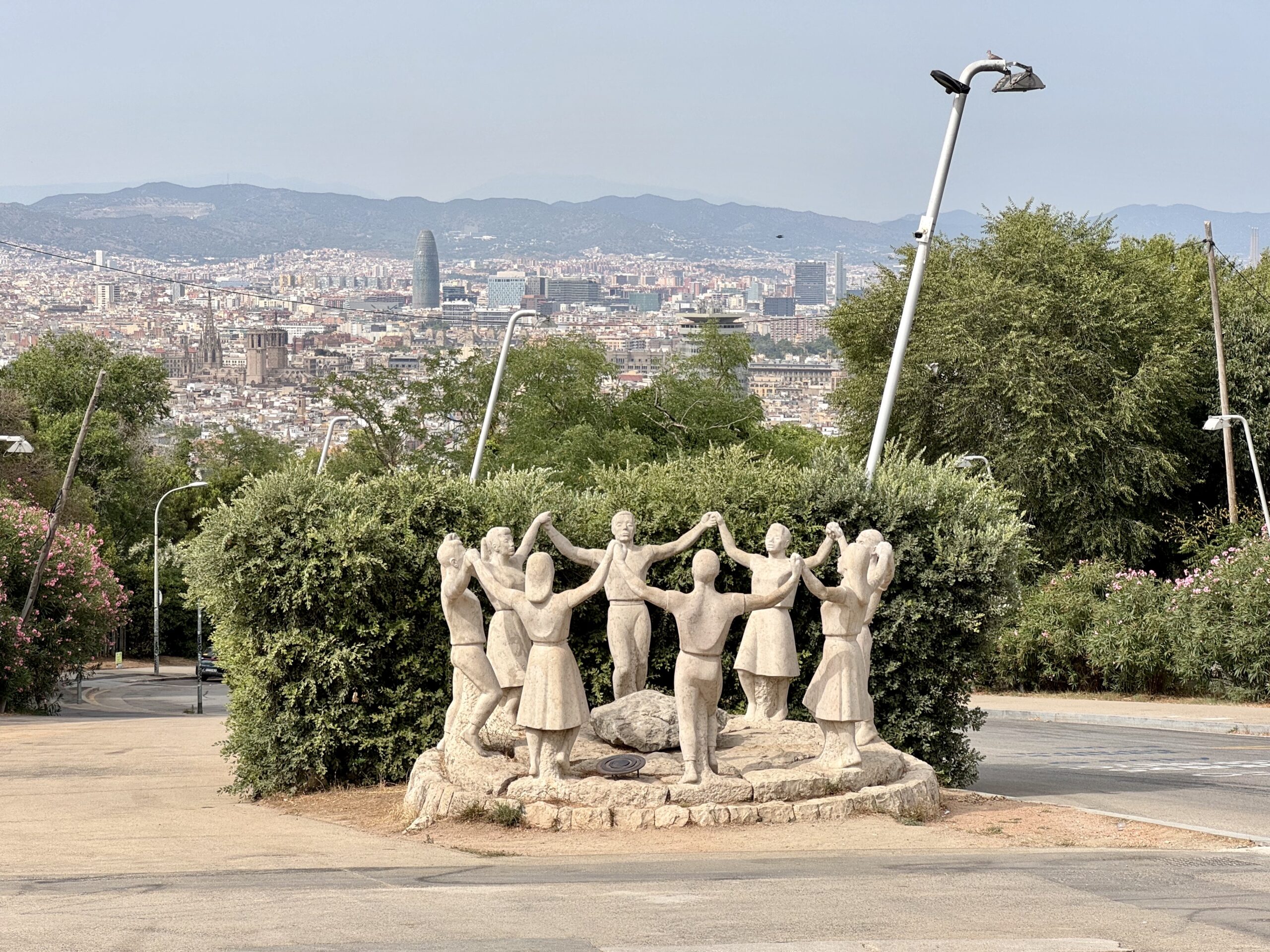 Stone sculpture of a circle of people holding hands on a pedestal in a park, with a city skyline and hills in the background.