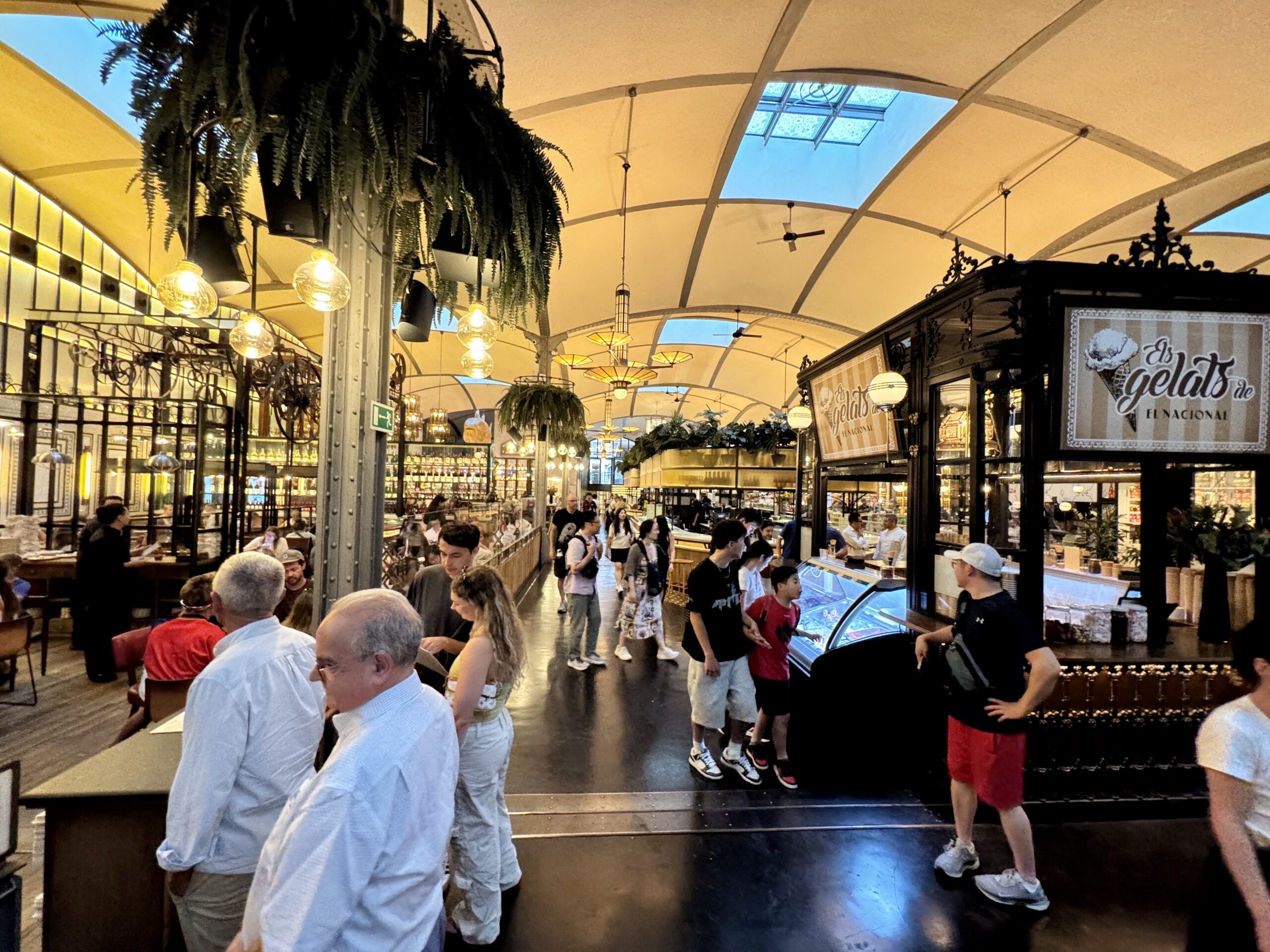 Busy indoor market with a gelato counter, people waiting, hanging lights, and hanging plants.