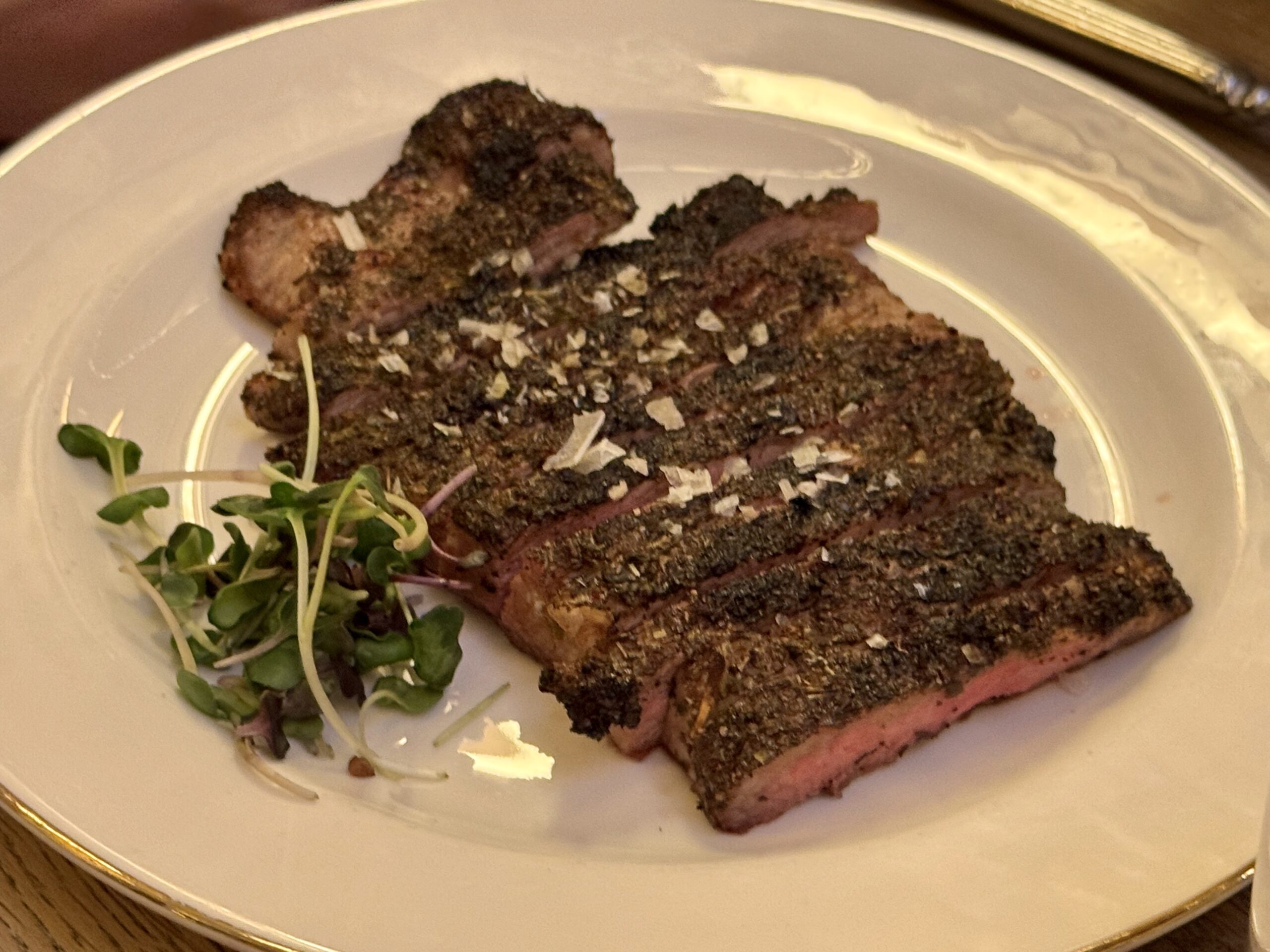 Sliced herb-crusted steak on a white plate with microgreens garnish on the side in a dining setting.