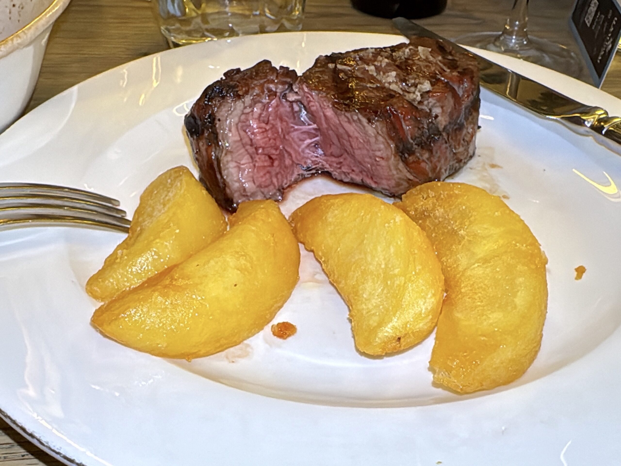 Thick slice of medium-rare steak with a pink center on a white plate, alongside several golden potato wedges.
