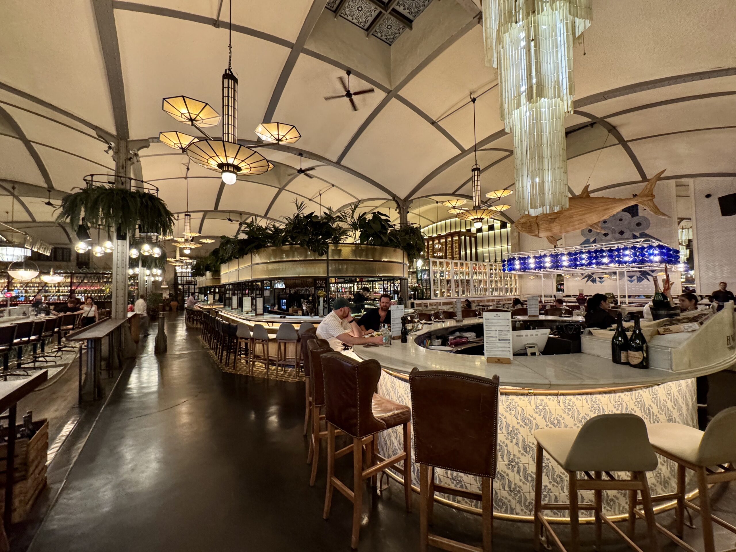 Indoor restaurant/bar with a curved, arched ceiling and warm pendant lights above a circular marble bar with stools outside seating areas nearby.