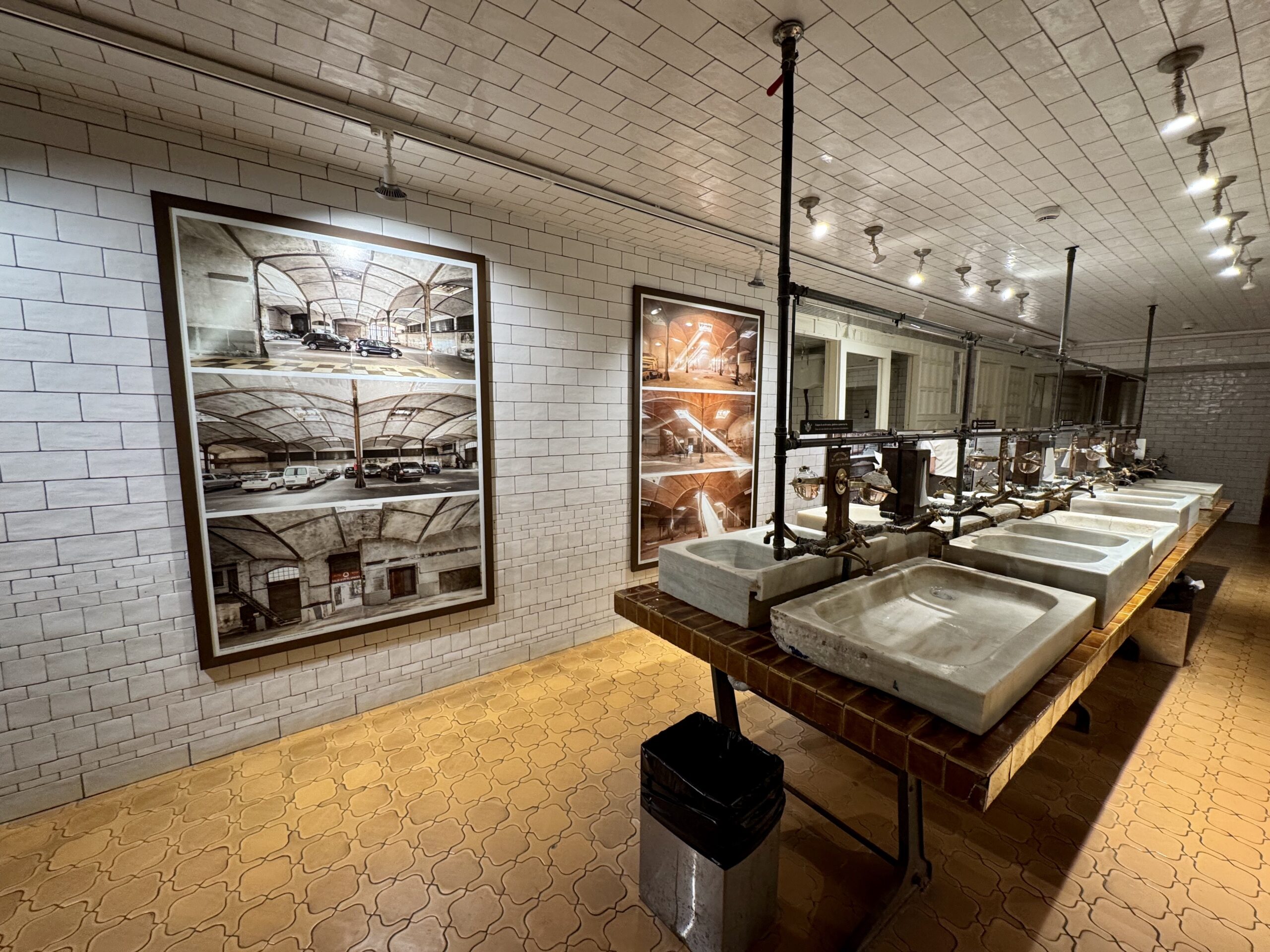 Interior museum or exhibit space with framed photographic panels along a tiled wall, and row of large concrete sinks connected to metal piping and fixtures above.