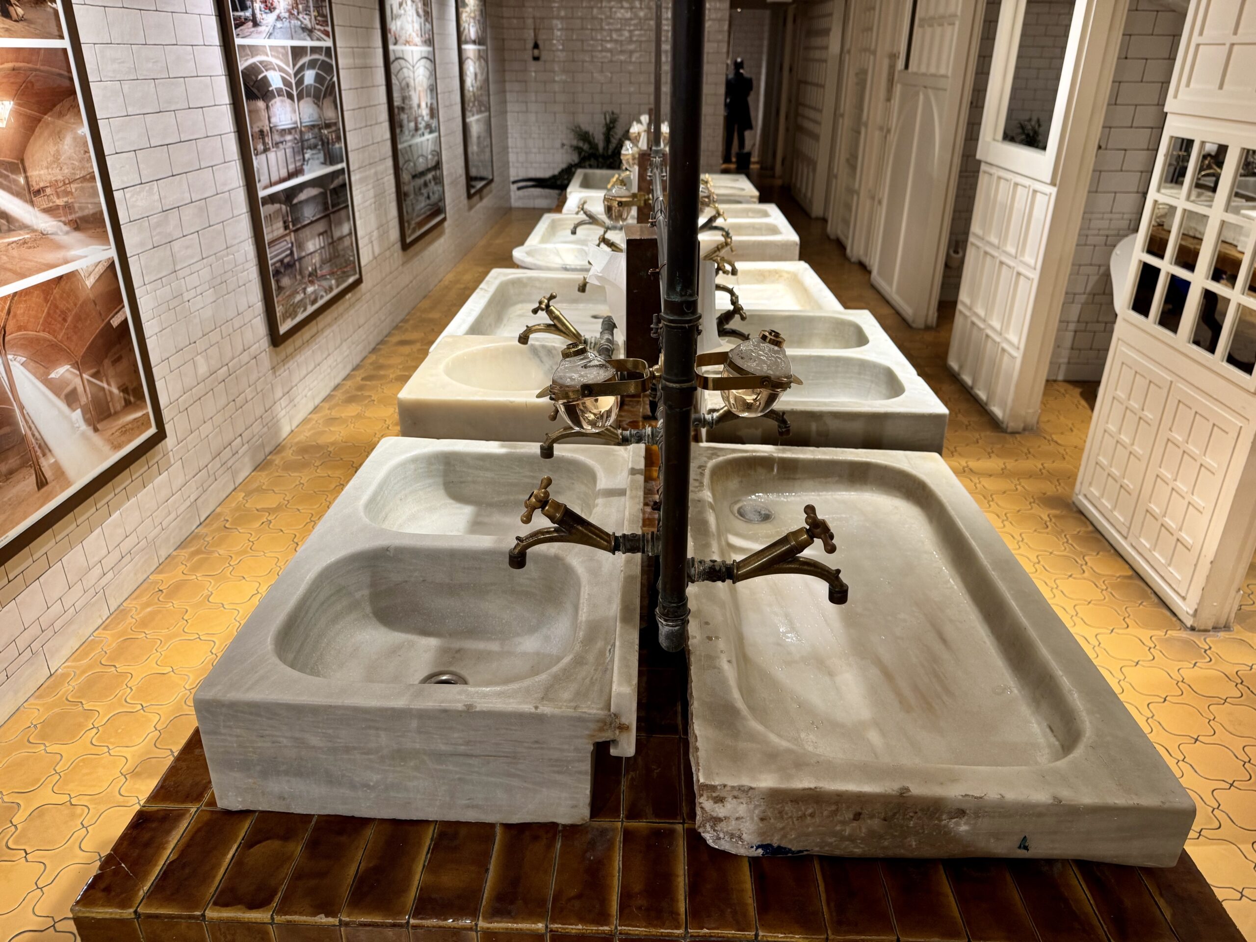 Row of vintage sinks with brass taps in a museum exhibit along tiled walls and a wall of framed photos far side.