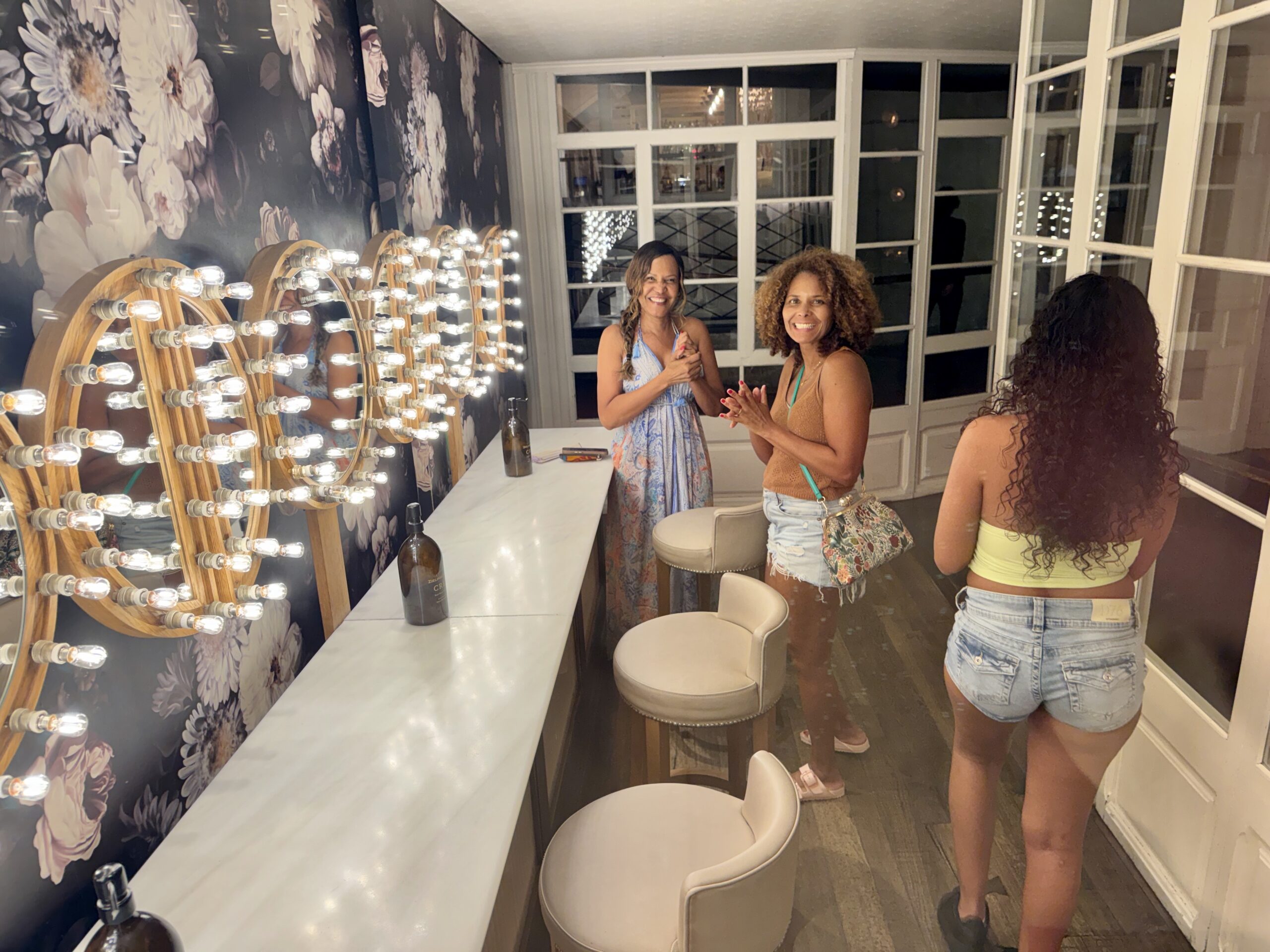 Two women smile at a makeup vanity with round light bulbs around a large mirror and floral wallpaper in the background.