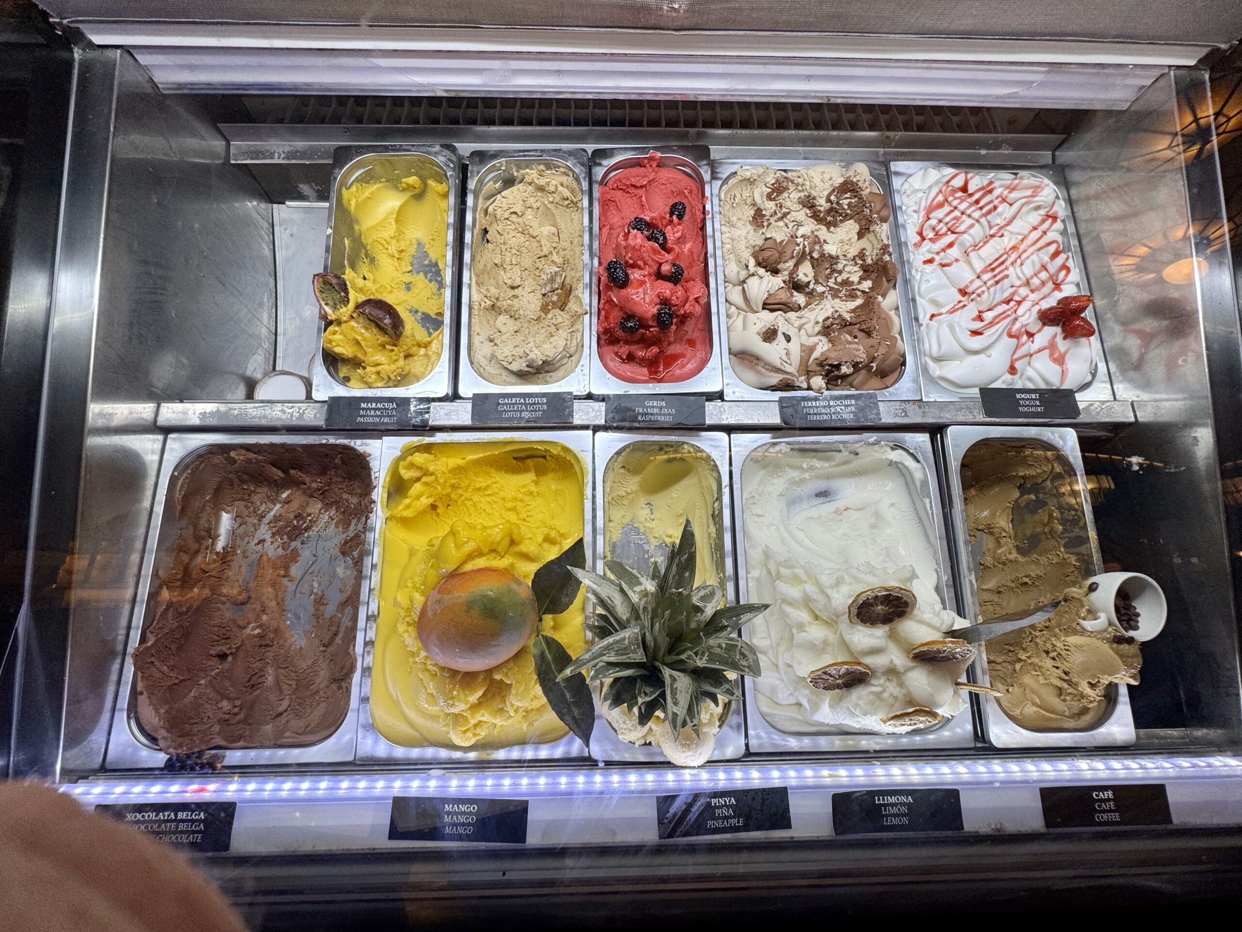 Row of gelato scoops in a display case: mango, passion fruit, berry sorbet, chocolate hazelnut, vanilla with a strawberry swirl visible.