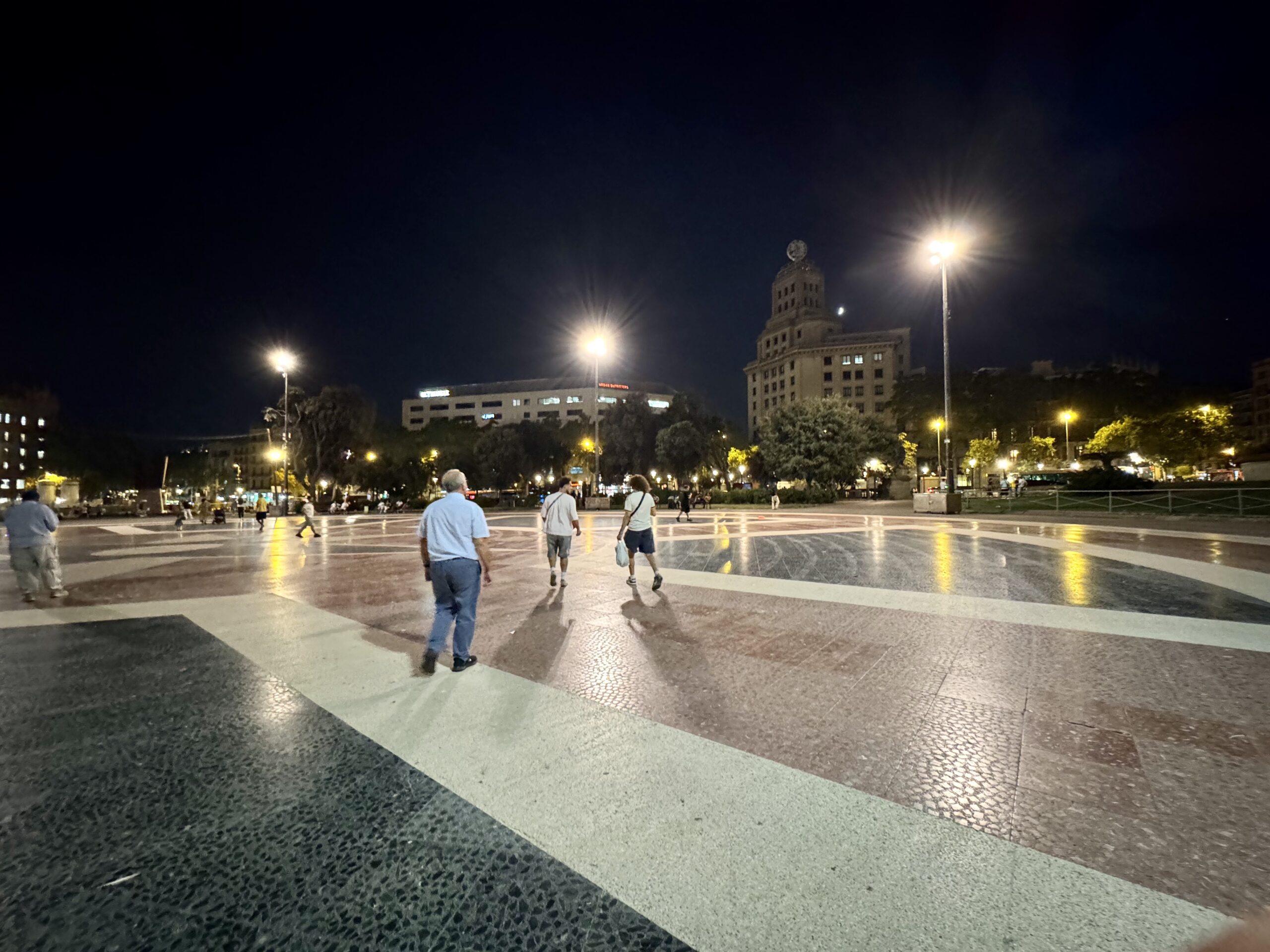 Night scene of a illuminated urban plaza with people walking, tall streetlights, and modern buildings in the background.