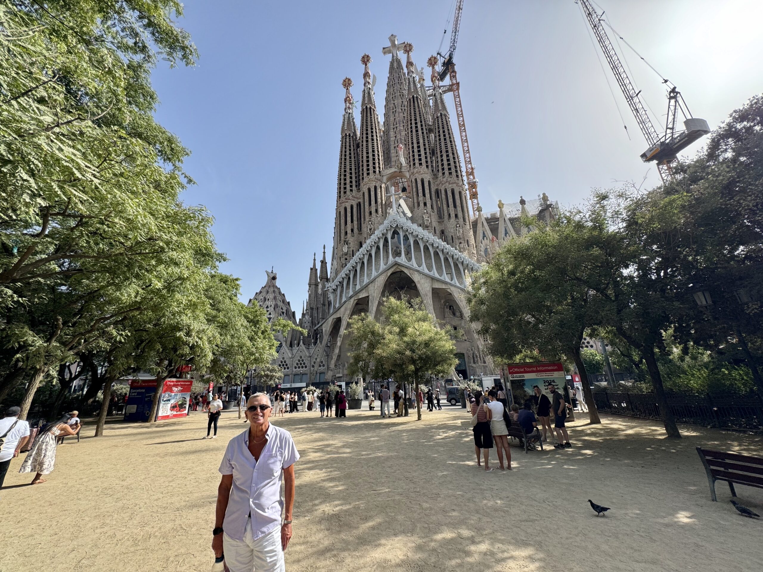 Sagrada Família in Barcelona with cranes above, visitors walking in the sunlit plaza surrounded by trees.