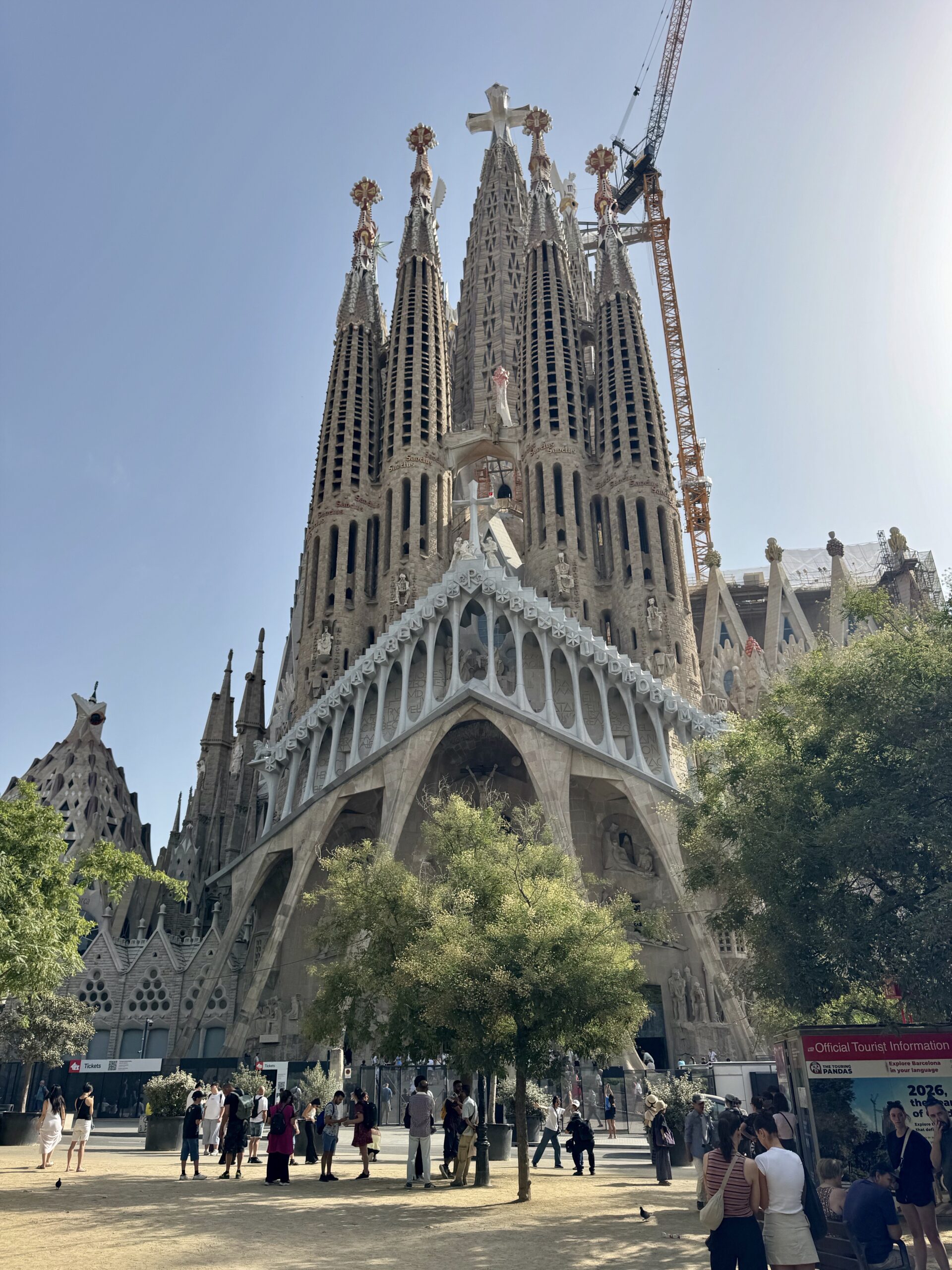 Front view of the Sagrada Família in Barcelona with tall spires and a construction crane against a clear blue sky, people gathered at the entrance