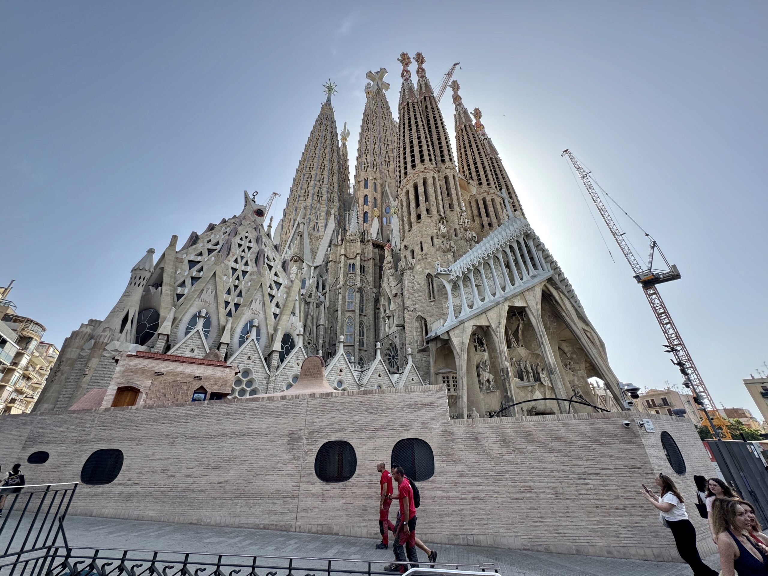 Sagrada Família in Barcelona under construction with tall spires and construction cranes against a clear blue sky, with pedestrians in the foreground.