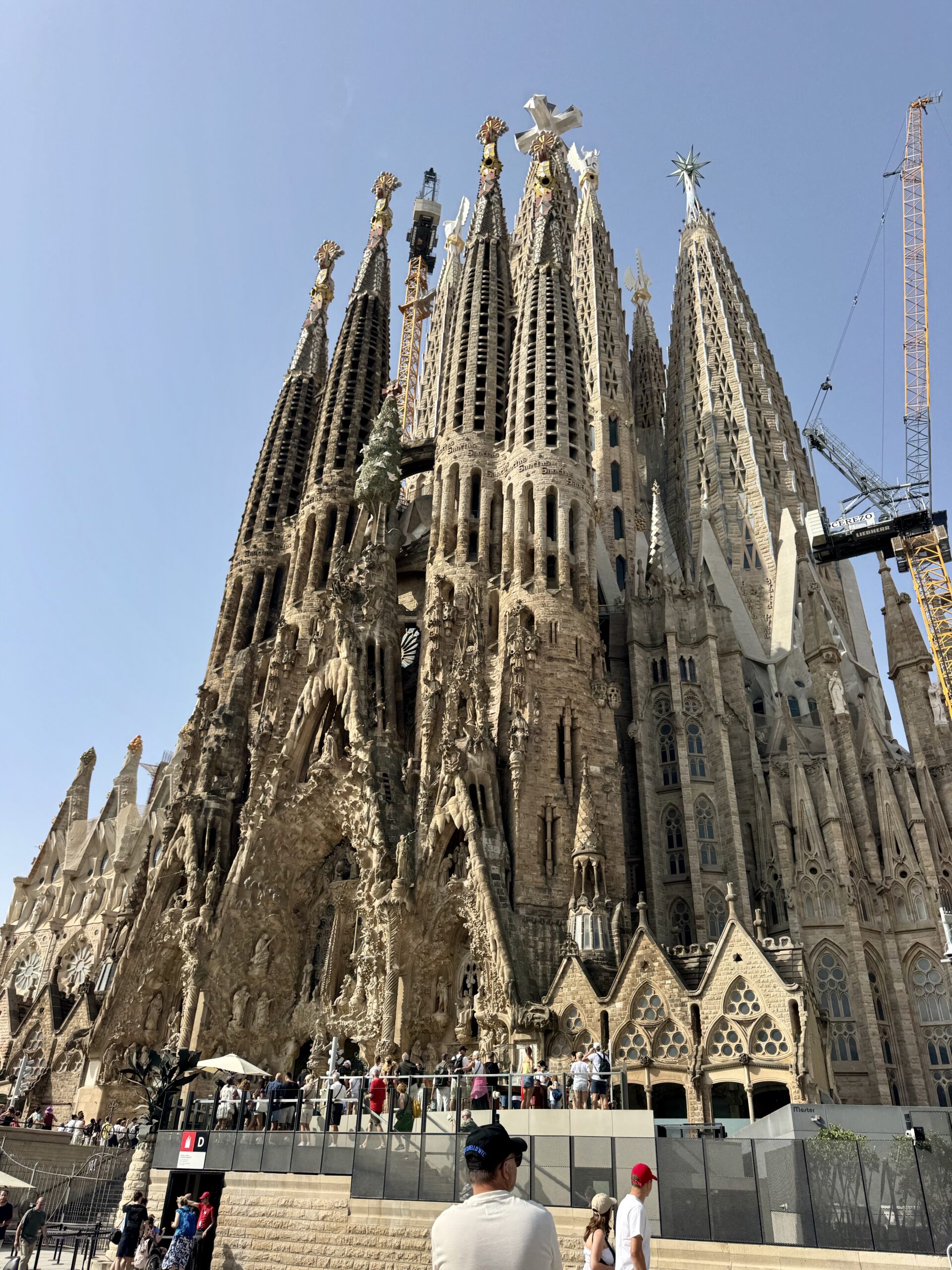 Sagrada Família's intricate spires under construction with cranes against a blue sky; visitors at the base viewing area.